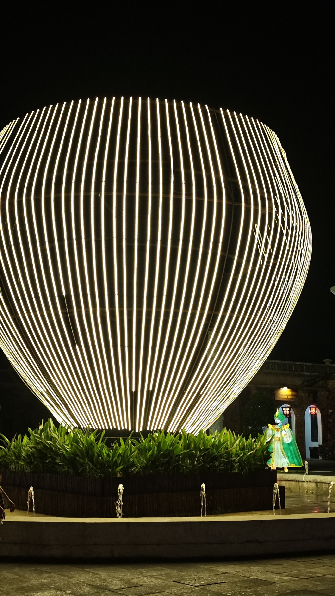 The photograph captures a nighttime scene in the city of Hội An Ward, Vietnam. The foreground features a decorative fountain enclosed by a low concrete wall. From the fountain's waters, several small streams of water shoot upwards. Above the fountain is a planting area with dark green, leafy plants in front of a short, dark-colored fence. Behind this planting area looms a large, illuminated structure constructed from evenly-spaced vertical, glowing lines, creating a rounded, almost egg-like shape. To the right of this structure stands a brightly colored, illuminated figure in green robes and gold accents. Further in the background, glimpses of buildings are visible, one featuring an arched doorway. The overall ambiance is illuminated and festive, contrasting with the deep black sky above.