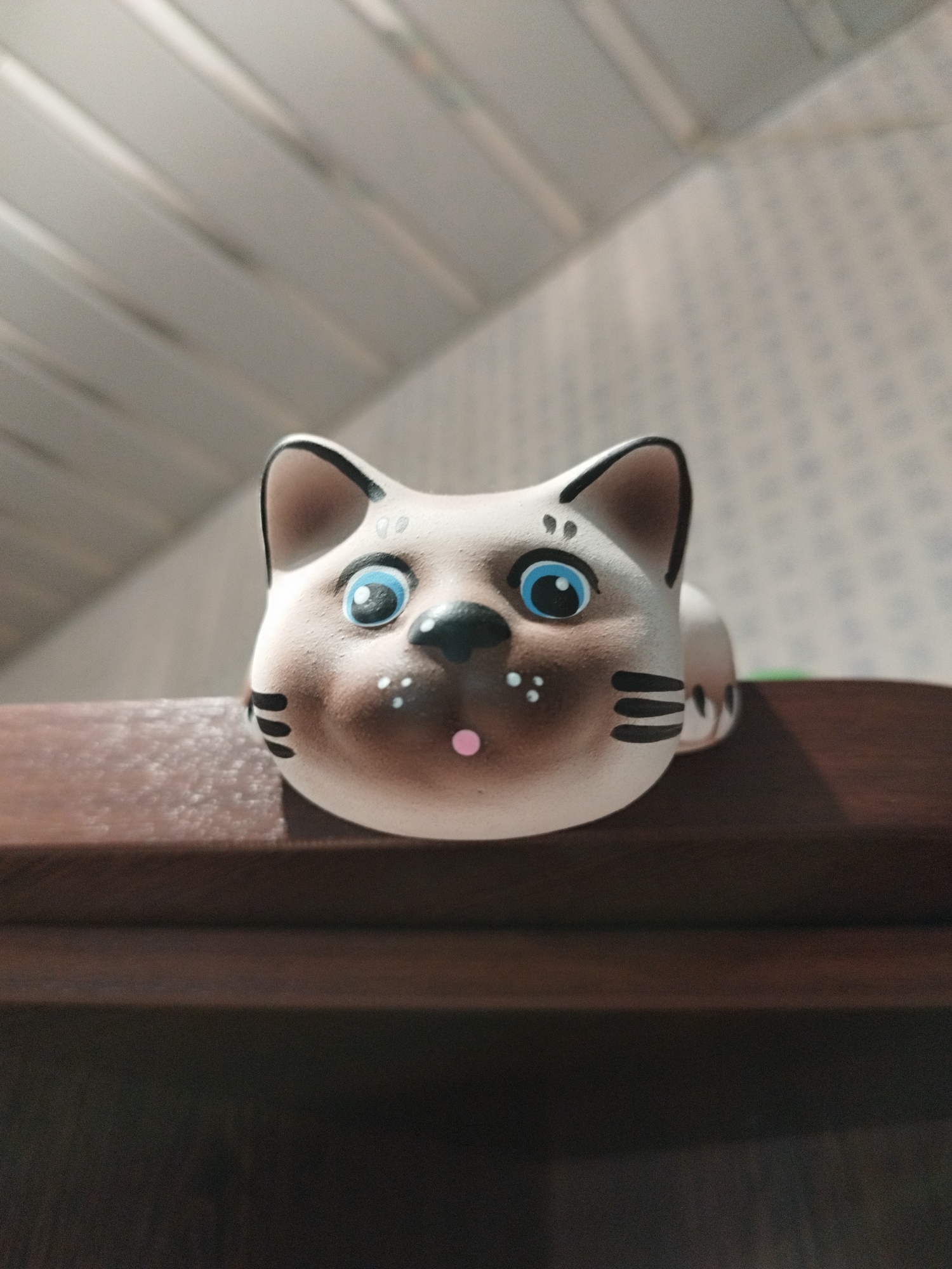 The image shows a close-up, eye-level, indoor shot of a decorative ceramic cat figurine positioned on a dark wooden surface. The figurine has a light brown head with large blue eyes, black stripes on its cheeks, and a small pink nose. It appears to be part of a larger object, possibly a planter or a bank, with its body mostly obscured. The background features a light-colored ceiling with a textured pattern and diagonal lines, suggesting a room interior. The lighting is soft, casting subtle shadows. The scene does not depict any specific location in the city of Saint Petersburg, Russia, as it is a generic indoor shot of an object.