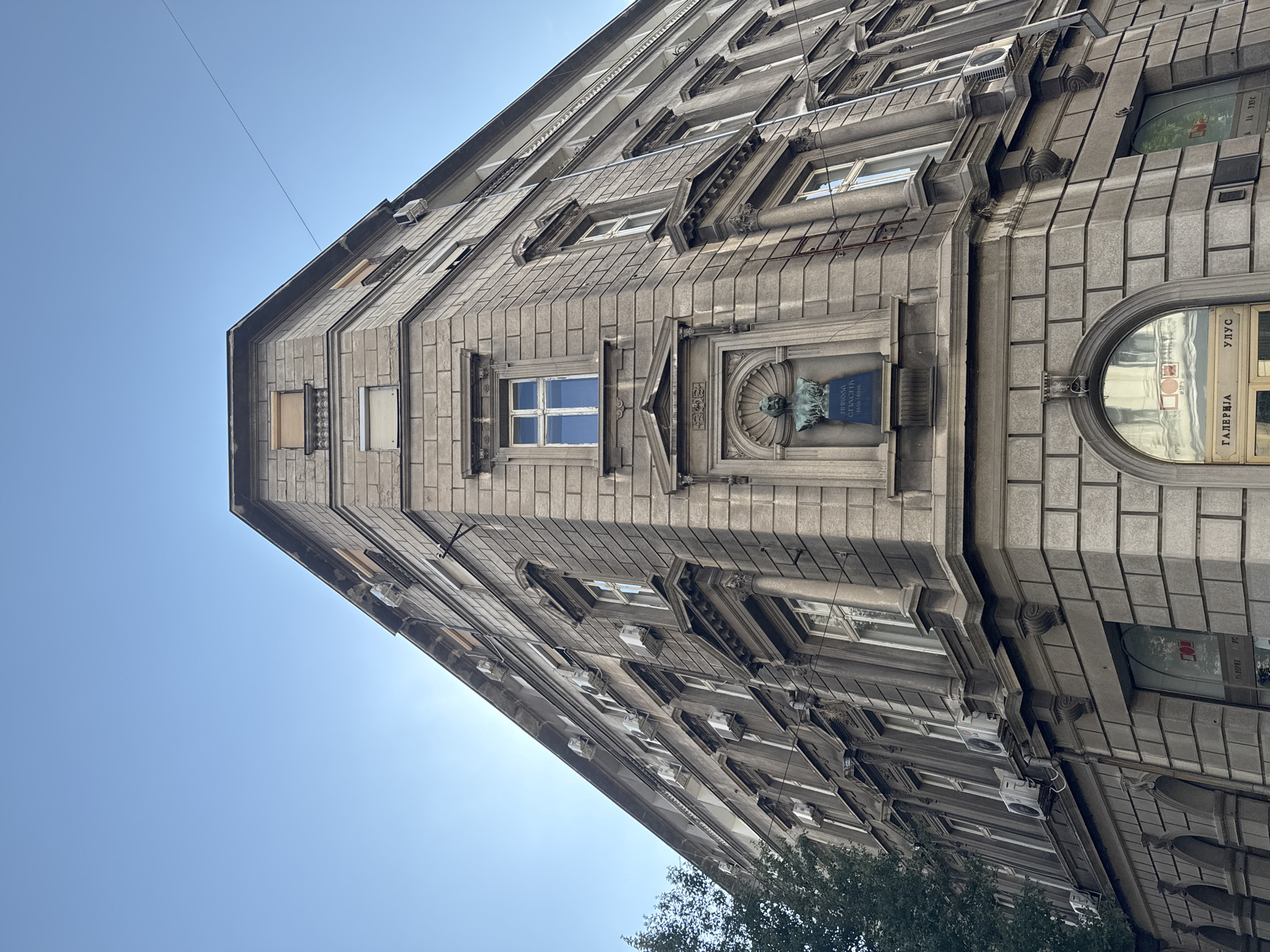 The image depicts a close-up, low-angle view of the exterior of a grey stone building in Belgrade, Serbia. The building has multiple stories, with numerous windows and air conditioning units visible on its facade. At the corner of the building, on the second story, is a bronze bust of a man in a niche, with a plaque below it inscribed with Cyrillic text. This bust is of Nikola Pašić, a prominent Serbian statesman. The building's architecture features decorative stone carvings around the windows and along the cornices. The sky above is clear and blue. A portion of a green tree is visible on the left side of the image. The building appears to be located in an urban setting, with hints of shop signs visible at the ground level.