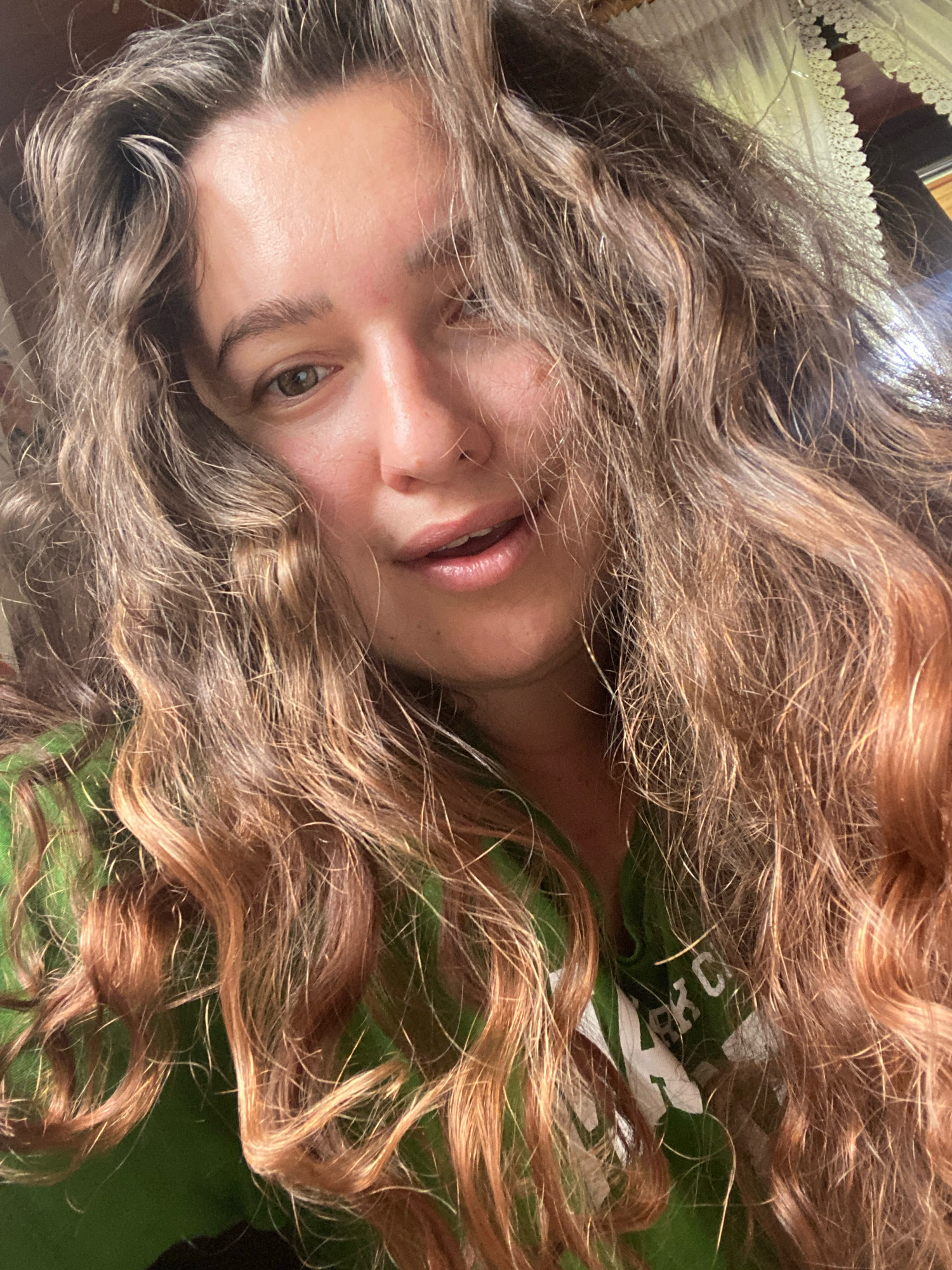 A close-up, front-facing selfie of a young woman with long, wavy, light brown hair, partially covering her face. She is wearing a green hooded sweatshirt. Her eyes are green, and her mouth is slightly open. The background is blurred, suggesting an indoor setting, possibly a home. There are no discernible landmarks or indicators that place the image in the city of Perm, Russia.