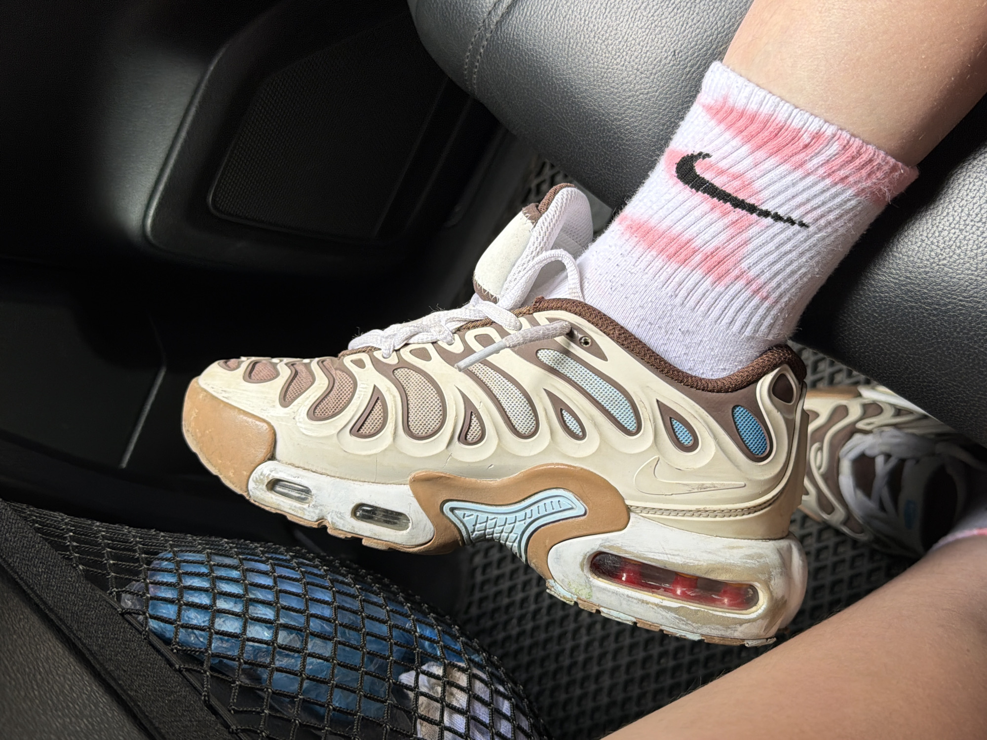 A close-up shot focuses on a person's legs and feet inside a vehicle, as indicated by the dark interior and car seat texture. The primary subject is a pair of beige and brown Nike Air Max Plus sneakers, worn with white crew socks that have a pink tie-dye pattern and the iconic black Nike swoosh logo. The shoes appear well-worn, with some scuff marks and dirt visible on the soles and white sections. The person's legs are bare from the knee down. In the foreground, a black mesh pocket is visible, containing a blue object, possibly a water bottle. The lighting suggests it might be daytime, possibly with natural light filtering into the car. The image seems to capture a casual moment, with no strong emotions conveyed. The setting appears to be inside a car, possibly on a trip given the inclusion of a mesh pocket often found in vehicles. The location context provided is Turbaza Katun', Russia, but this is not directly discernible from the visual elements of the image itself.