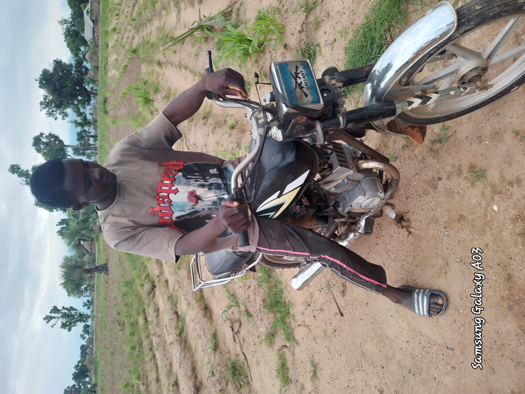 A young Black male sits on a motorcycle in a rural area. He is wearing a light brown t-shirt with a graphic design on it, dark pants with pink stripes, and grey flip-flops. The motorcycle is black and silver with a chrome exhaust pipe and handlebars. The background shows a field with rows of plants, trees, and huts, suggesting a village setting in Angwan Hausawa, Nigeria. The sky is overcast. The Samsung Galaxy A03 watermark is visible in the bottom left corner of the image.