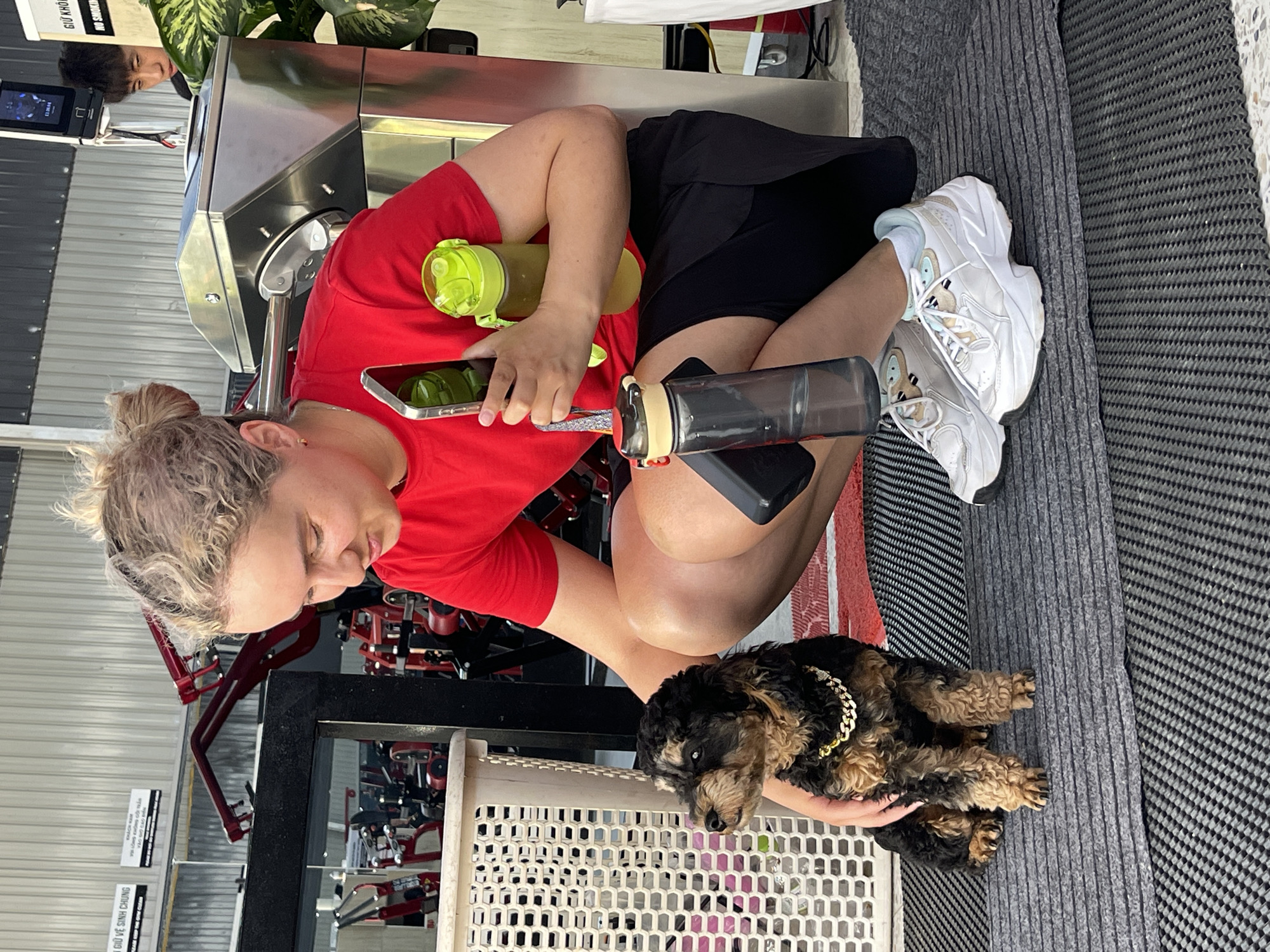 A woman is seated in a gym, holding a black and brown dachshund dog with a gold chain around its neck. The woman is wearing a red t-shirt and black shorts, with white sneakers on her feet. She is holding a smartphone in her left hand and a black water bottle in her right hand, with a green water bottle resting beside her. The dog is held over a white plastic basket. In the background are various gym machines and a mirrored wall reflecting the gym equipment. A sign is visible in Vietnamese. The gym appears to be located in Da Lat, Vietnam.