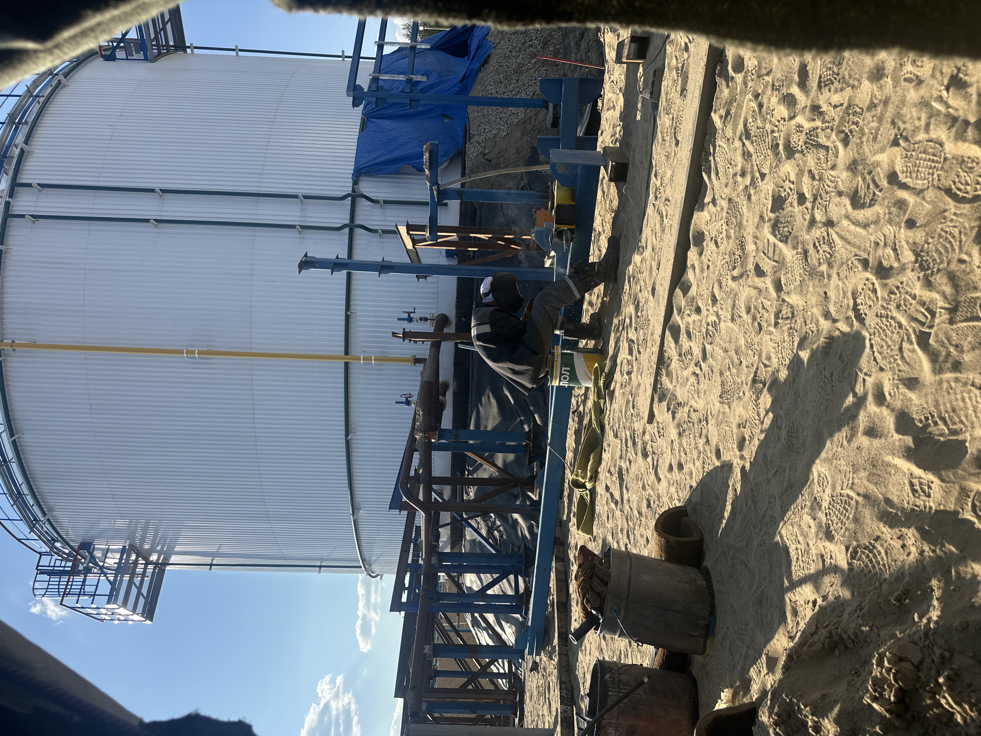 A worker in a grey jumpsuit and white hard hat is welding in front of a large white cylindrical tank in Чикшино, Russia. The tank has a grated walkway and ladder on its left side and green pipes running vertically along its surface. A blue tarp is draped over some construction materials to the right of the tank. The ground is covered in sand with numerous footprints. Several buckets, gloves, and rolled-up materials are scattered on the sand in the foreground. The scene appears to be a construction or industrial site under a clear blue sky with a few clouds.