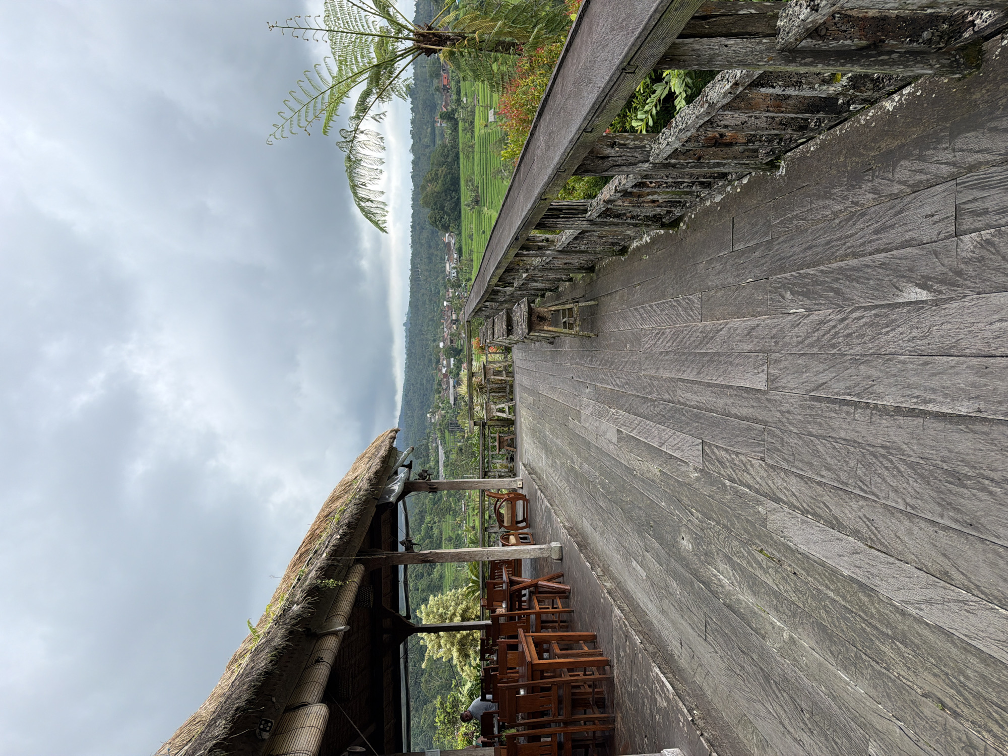 The image captures an elevated wooden walkway in an unidentified location within the city of Unknown, Indonesia. The walkway is constructed of weathered, graying wood planks, leading away from the viewer at a sharp perspective. A rustic wooden railing borders the walkway. Beyond the railing, a vibrant green landscape stretches out, featuring rice paddies and distant structures nestled amidst dense foliage.
To the left of the walkway, a partially visible structure with a thatched roof provides shade and shelter. Wooden chairs and tables are arranged beneath this structure, indicating a possible dining or seating area. An individual is visible in this area, seated at one of the tables. The sky above is overcast with thick, gray clouds. A tall fern-like plant is positioned near the edge of the walkway.