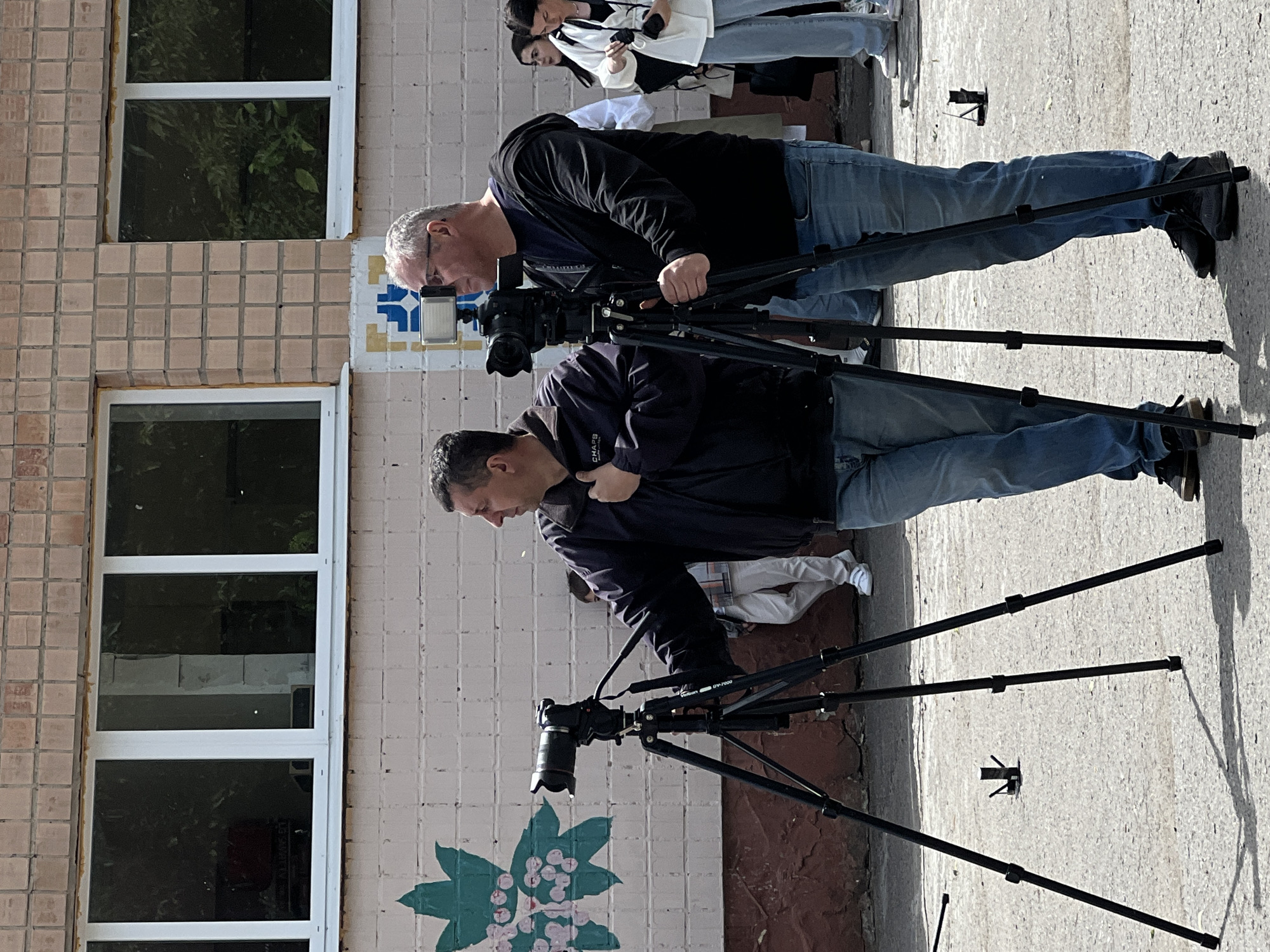 Two men stand on a sidewalk, each operating a camera mounted on a tripod. They face a building with tiled walls and several windows. The man closer to the camera wears a dark jacket and jeans; the other man wears a black shirt and jeans. Behind them are several other people. A green and pink floral design is painted on the wall near the base of the building. The location is in the city of Ternopil, Ukraine.