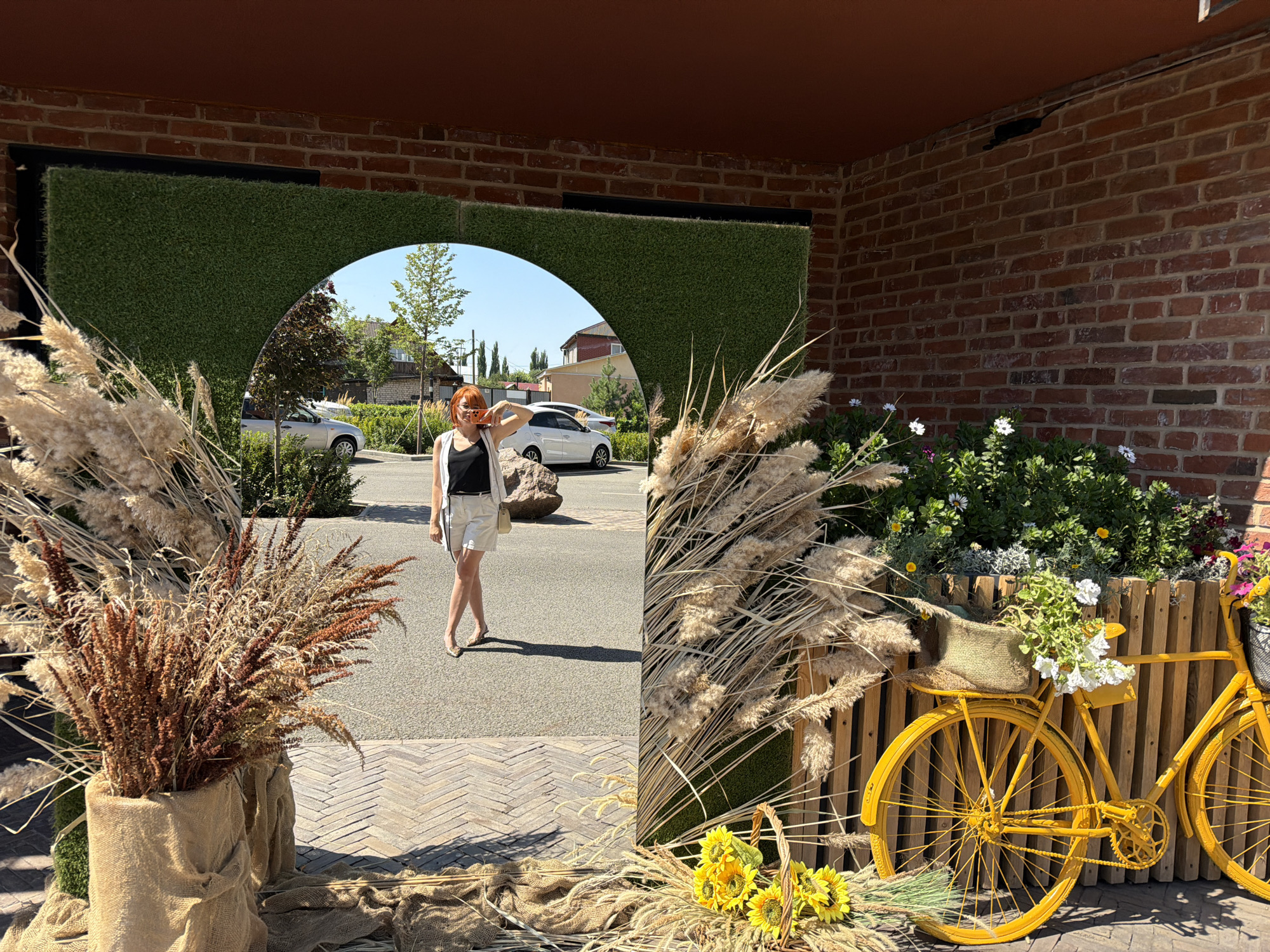 The image captures a street scene in Buzuluk, Russia, featuring an outdoor area decorated with dried grasses and flowers, and a bright yellow bicycle. A woman with reddish-orange hair, wearing a black tank top and white shorts, is taking a selfie with her phone, reflected in a large mirror. She is standing in what appears to be an archway covered in artificial grass. In the background, cars and buildings are visible. To the right of the archway, a yellow bicycle is adorned with a burlap bag and white flowers. The overall setting suggests a decorative outdoor space, possibly part of a cafe or public area.