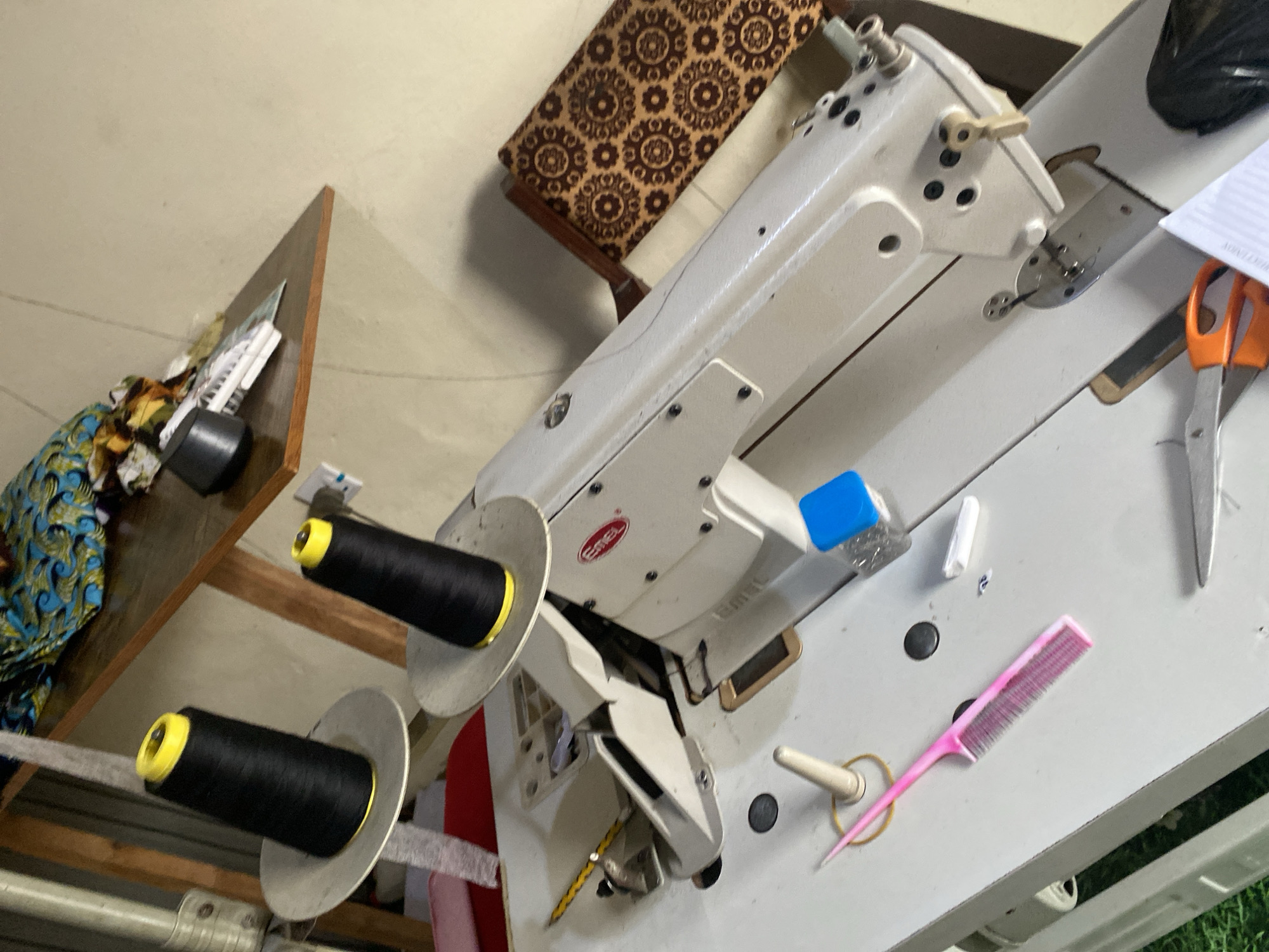 The image depicts a sewing machine in a workshop located in Jauro Yinu, Nigeria. The sewing machine is a light-colored industrial model with the brand name "Eemel" visible on its side. Two large spools of black thread with yellow tops are mounted on a spindle above the machine. Various sewing tools and materials are scattered on the white surface of the sewing machine table, including a pink comb, a white thimble, a blue container, a white chalk, and a pair of orange-handled scissors. In the background, a wooden shelf holds a piece of colorful fabric. The workshop environment appears functional, with the tools and equipment suggesting ongoing sewing activities.