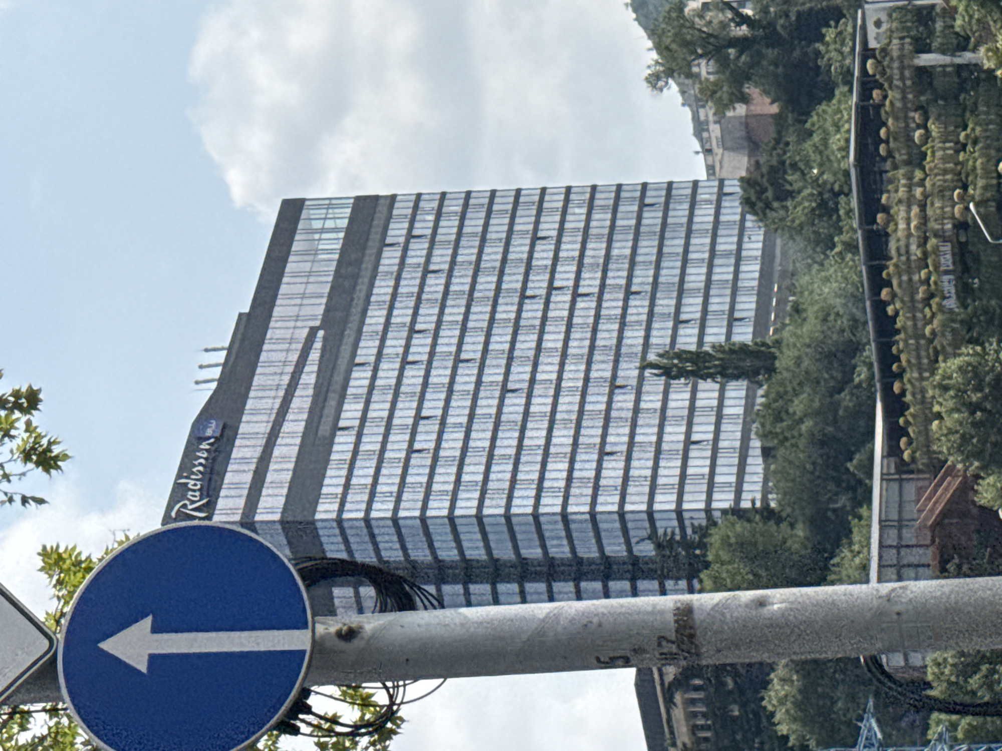 A large, modern, glass-fronted building with the Radisson Blu logo prominently displayed on its facade stands tall against a cloudy blue sky. In the foreground, a grey metal pole holds a circular blue traffic sign with a white upward-pointing arrow, indicating a one-way street. This scene is situated in Tbilisi, Georgia, with lush green trees and vegetation surrounding the building, suggesting an urban or semi-urban environment. Other structures, including what appears to be a multi-level building with outdoor terraces and staircases, are visible behind the main building and amidst the greenery. The overall impression is of a contemporary cityscape integrated with natural elements.