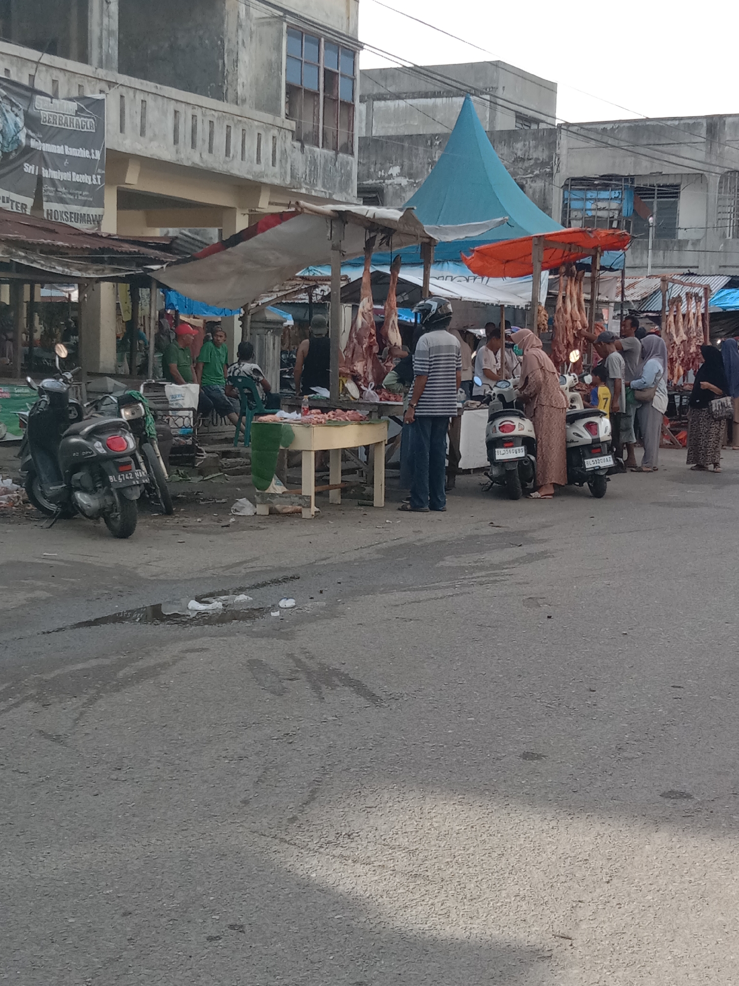The image depicts a bustling meat market scene, likely in the city of Lhokseumawe, Indonesia. Pieces of meat are hanging from the makeshift stalls under various canopies, and a crowd of people is gathered around a table where meat is being sold. Several individuals are wearing traditional Muslim attire, including a woman in a hijab. A man in a striped shirt stands near a scooter with the license plate BL3560KBS. Two other scooters are also parked nearby, one with license plate BL4742 NAK and the other with BL592QUAZ. A group of men in green shirts are sitting further in the background. The overall environment appears to be a somewhat cluttered public space with buildings in the background. The pavement is stained, suggesting high foot traffic and potential spills. The image conveys a sense of a lively, local marketplace in the city.