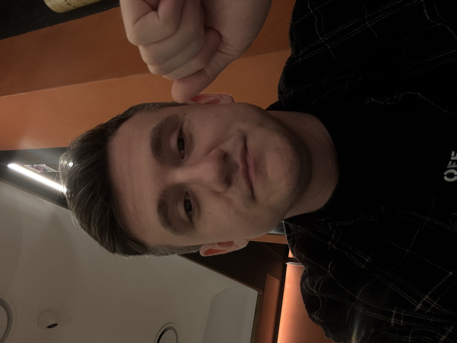 The image displays a close-up indoor portrait of a male individual, approximately 20-30 years old, situated in a location within Verona, Italy. The subject has short, dark brown hair and a neutral to slightly smirking facial expression, looking directly at the camera. He is wearing a dark plaid button-down shirt over a black t-shirt, with a white logo partially visible on the t-shirt. The individual's right hand is raised to the level of his right ear, clenched into a fist. The background elements include a light-colored ceiling with a recessed circular light fixture and a small dome-shaped security camera (CCTV). A vertical strip light illuminates a dark wall or structural element, adjacent to an orange-brown vertical panel. A bottle, possibly a wine bottle with text, is partially visible against a dark background on the far right. The scene is illuminated by artificial lighting, creating a somewhat dim overall ambiance.