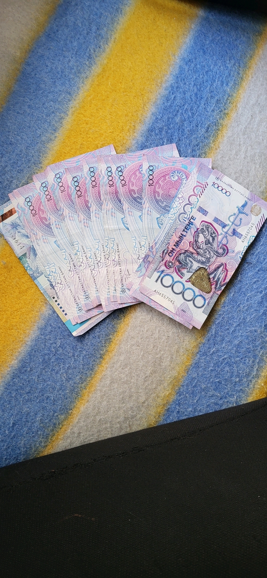 A fan of ten 10,000 tenge banknotes lies on a blue, yellow, and white striped blanket. The banknotes are oriented diagonally from the lower left to the upper right of the frame, with the top banknote slightly overlapping the others. Each banknote is predominantly pink and blue, with intricate patterns and the denomination "10000" clearly visible on most of them. A small, gold-colored coin is placed on the top banknote, obscuring part of its design. The background is a soft, textured blanket. The lower portion of the image shows a dark, plain fabric surface, possibly a piece of clothing or furniture. The scene is indoors, likely in a domestic setting.