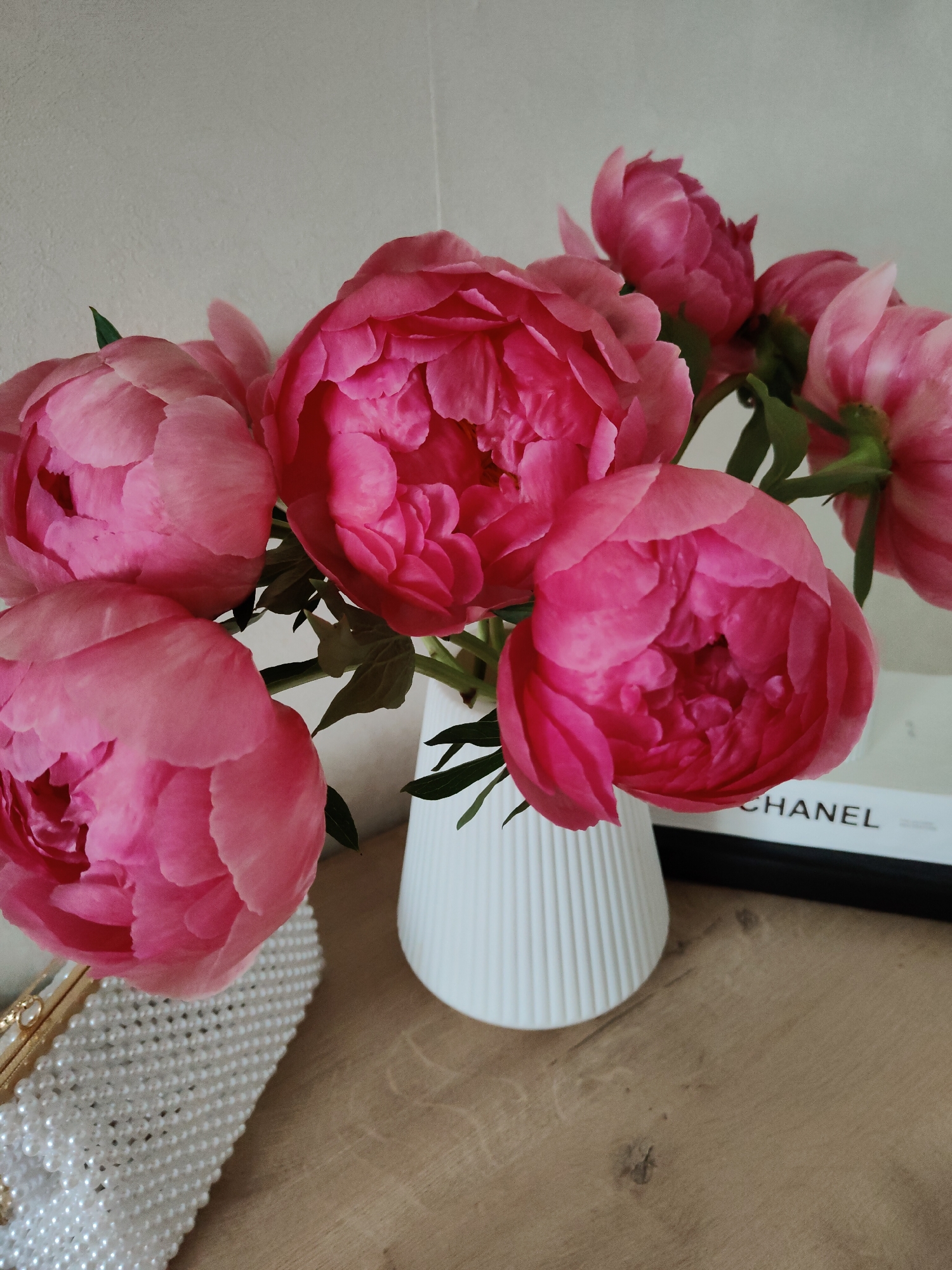 An indoor close-up shot captures a bouquet of vibrant pink peonies arranged in a white, vertically ribbed cylindrical vase. The flowers, in various stages of bloom, display deep fuchsia and lighter pink petals, with visible green stems and leaves. The vase is positioned on a light-colored wooden surface that shows grain patterns and a few darker marks. To the left foreground, partially visible, is a white object with a textured surface composed of numerous small, spherical elements, resembling a pearl-beaded clutch or bag, with a metallic gold-toned clasp. To the right background, behind the vase and some flowers, a rectangular object, possibly a book or decorative box, is visible with the word "CHANEL" printed in black capital letters on its side. The background is a plain, light grey wall. The scene is located in an unspecified indoor setting; no city or geographical location is discernible.