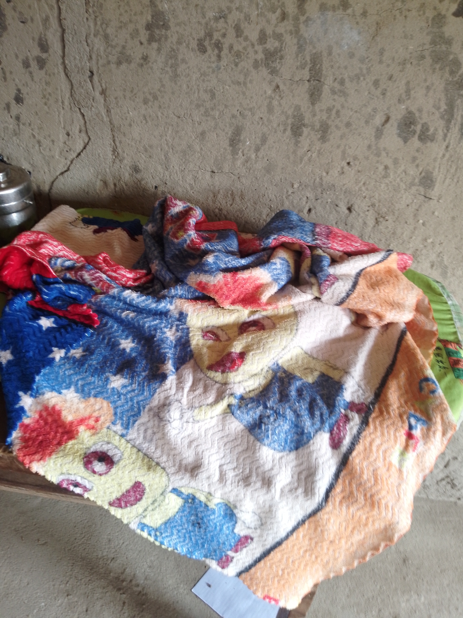 A folded patterned blanket is displayed on a surface, possibly a mat, against a rough concrete wall. The blanket features a colorful design with cartoon characters, stars, and abstract patterns in shades of blue, red, yellow, orange, and white. The scene is indoors, with a metallic object, possibly a lunchbox, visible on the left side. The lighting suggests an overcast environment. The location is Serti, Nigeria.