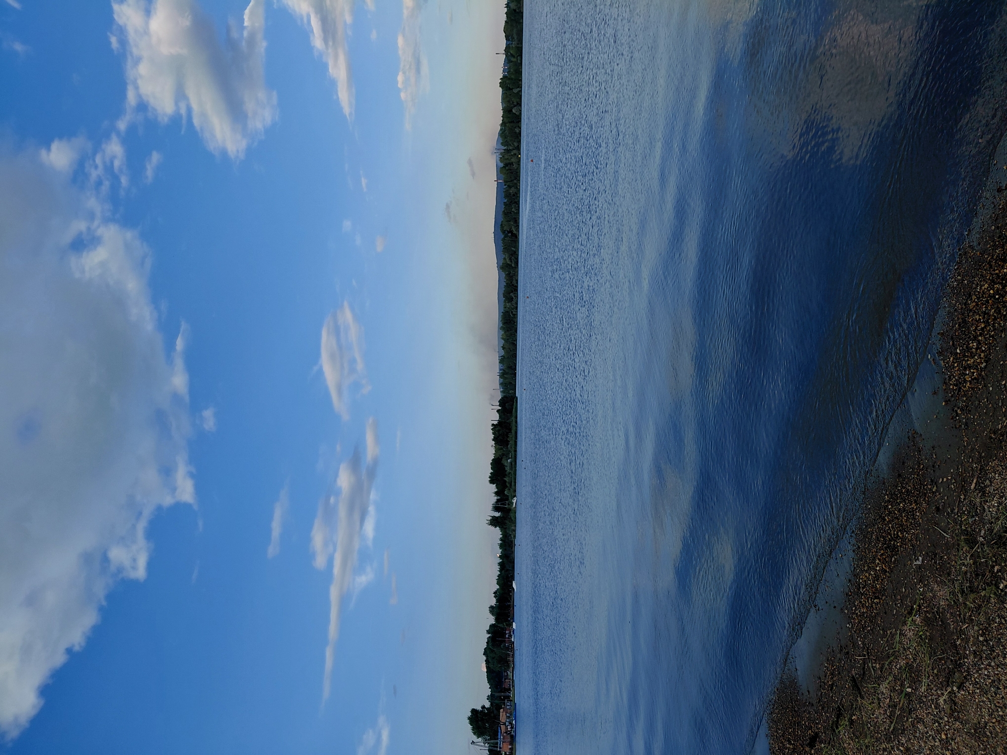 The image depicts a serene body of water, likely a river or lake, under a partly cloudy blue sky. The foreground features a coarse, pebbly shoreline. In the mid-ground, the water surface is calm with gentle ripples, reflecting the sky and clouds. Along the far shore, a dense line of trees forms a natural barrier, interrupted in places by what appear to be industrial structures and chimneys, suggesting an urban or industrial fringe. Several small boats and buoys are visible scattered across the water, indicating recreational or navigational activity. The overall scene conveys a tranquil yet somewhat industrial landscape, characteristic of the outskirts of a city like Magnitogorsk, Russia, which is known for its heavy industry and proximity to natural bodies of water.