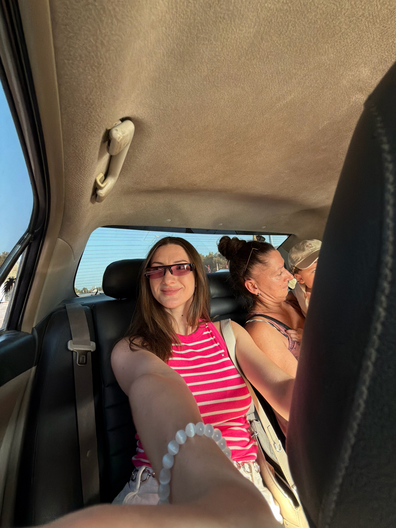 Three individuals, two adult females and one child, are seated in the back of a vehicle, likely a car or van. The front-most person, a woman wearing pink-striped top and sunglasses, is in the foreground, looking towards the camera. Behind her, another woman with her hair in a bun, wearing a patterned top, is looking to her right, facing away from the camera. A child wearing a cap is seated next to her, partially obscured. The interior of the vehicle appears to be upholstered in dark fabric, with a beige headliner. A seatbelt is visible. Through the rear window, a bright blue sky and some vegetation are discernible, suggesting an outdoor, possibly sunny environment. The scene is consistent with a journey within or near Sharm El Sheikh, Egypt, given the bright, clear sky and the presence of vegetation typical of the region.