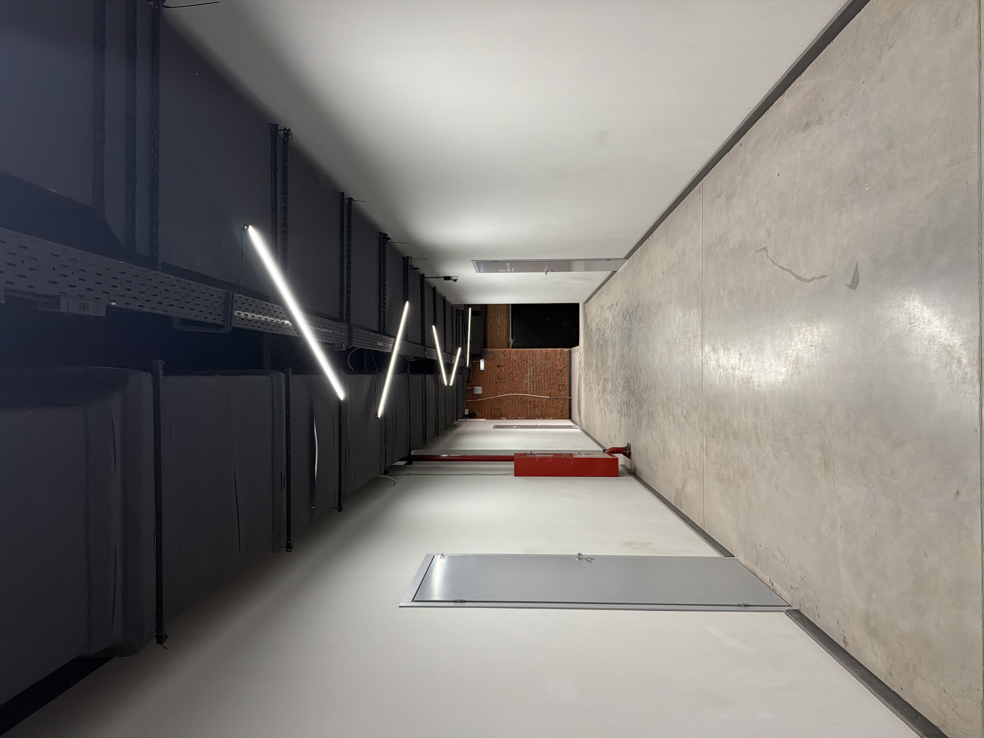 The image shows a stark, indoor corridor with industrial elements. The left wall features dark, vertical structures, possibly ventilation ducts, accompanied by bright, linear lights. The opposite wall is a light gray concrete surface. The floor is a flat, white plane, with large, gray access doors recessed within it. Further down the corridor, a red fire suppression system component is visible. At the end of the corridor, a brick wall is visible. The absence of people and the industrial aesthetic suggest a service area or back-of-house location, possibly in Saint Petersburg, Russia.