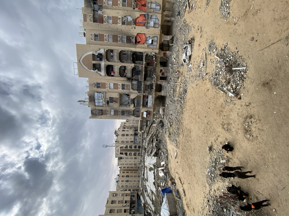 The image depicts an extensively damaged urban residential area in Khan Yunis, Palestinian Territory, under an overcast sky. The foreground features a large expanse of sandy ground littered with concrete rubble, twisted metal, and assorted debris. In the lower-left quadrant, a group of approximately six to seven individuals, primarily dressed in dark civilian clothing, are gathered; some stand, while others crouch on the sand. One individual wears an orange-colored vest. To the mid-right, a heavily damaged, inverted white vehicle wreck rests on the sand.
The mid-ground reveals significant destruction, with large piles of collapsed building materials, including concrete slabs and rebar. To the left, a makeshift shelter composed of a blue tarp and white canvas is visible, near which a person in a blue garment stands.
The background is dominated by multiple multi-story apartment buildings, uniformly constructed with light-colored facades. These structures exhibit severe damage: numerous shattered windows, missing wall sections exposing interiors, visible scorch marks, and widespread bullet or shrapnel impacts across their surfaces. Several balconies are partially destroyed or laden with debris and personal belongings. Rooftops display satellite dishes and telecommunication antennas. The building on the immediate right has prominent orange tarps covering lower-level damaged sections. The overall scene conveys widespread structural devastation.
