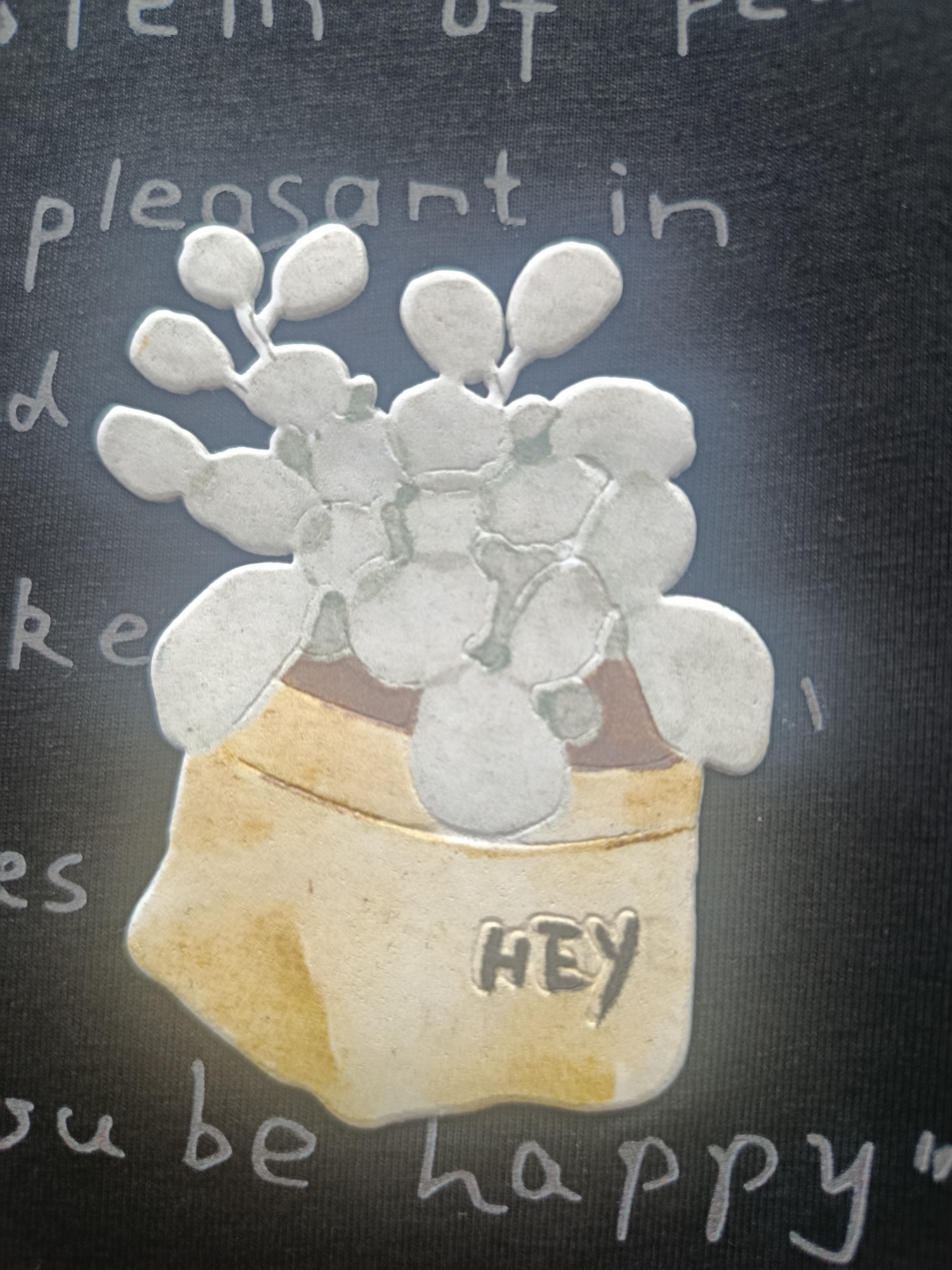The image is a close-up, angled shot of a dark, textured fabric with white lettering and a raised, three-dimensional metallic-looking artwork. The artwork depicts a potted plant with round, bulbous leaves, rendered in silver and brown tones. The pot is a pale yellow with brown accents. The word "HEY" is embossed on the pot. Above the plant, faint white lettering appears to read "stem of pe" and "pleasant in". Below the pot, more white lettering is visible, with "es" on the left and "ou be happy" on the bottom. There are no people or animals in the image. The environment is not discernible beyond the fabric background. This image is not related to Sertolovo, Russia.