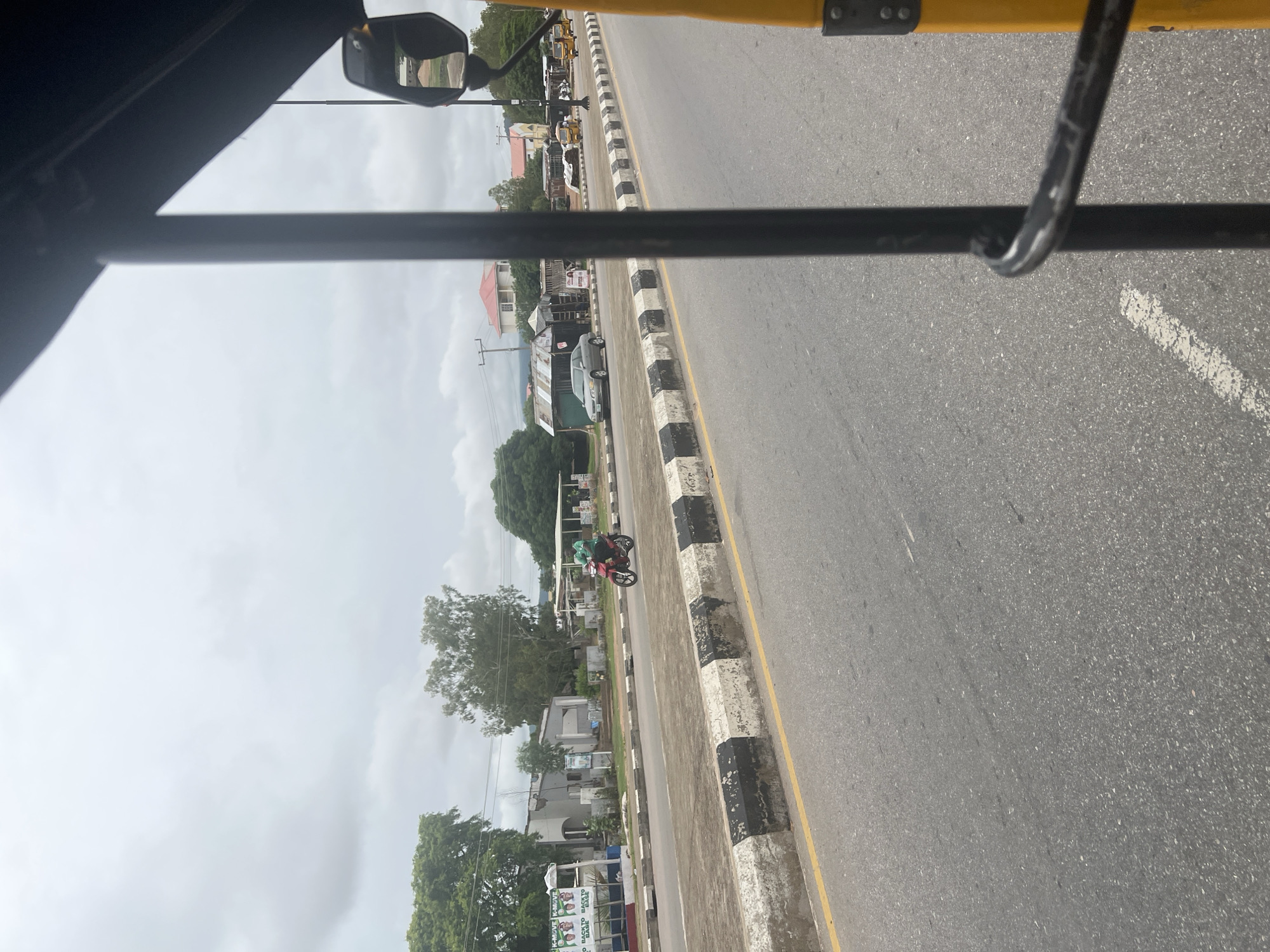 The image, taken from within a yellow vehicle, shows a street scene in Jalingo, Nigeria. The road is paved, with a distinct yellow line marking the edge of the lane and a concrete divider with black and white stripes separating the lanes. On the left side of the road, there are buildings and trees. One building has posters on its facade, and another appears to be under construction or in disrepair. A motorcycle with a rider wearing a green shirt is traveling on the road. Further down the street, a silver car is parked, and beyond that, several yellow motorized rickshaws (Keke Napep) are visible. The sky is overcast with grey clouds. The foreground is dominated by the interior of the vehicle, with a black pole and a black metal bar visible. A side mirror reflecting a building and trees is attached to the yellow vehicle.