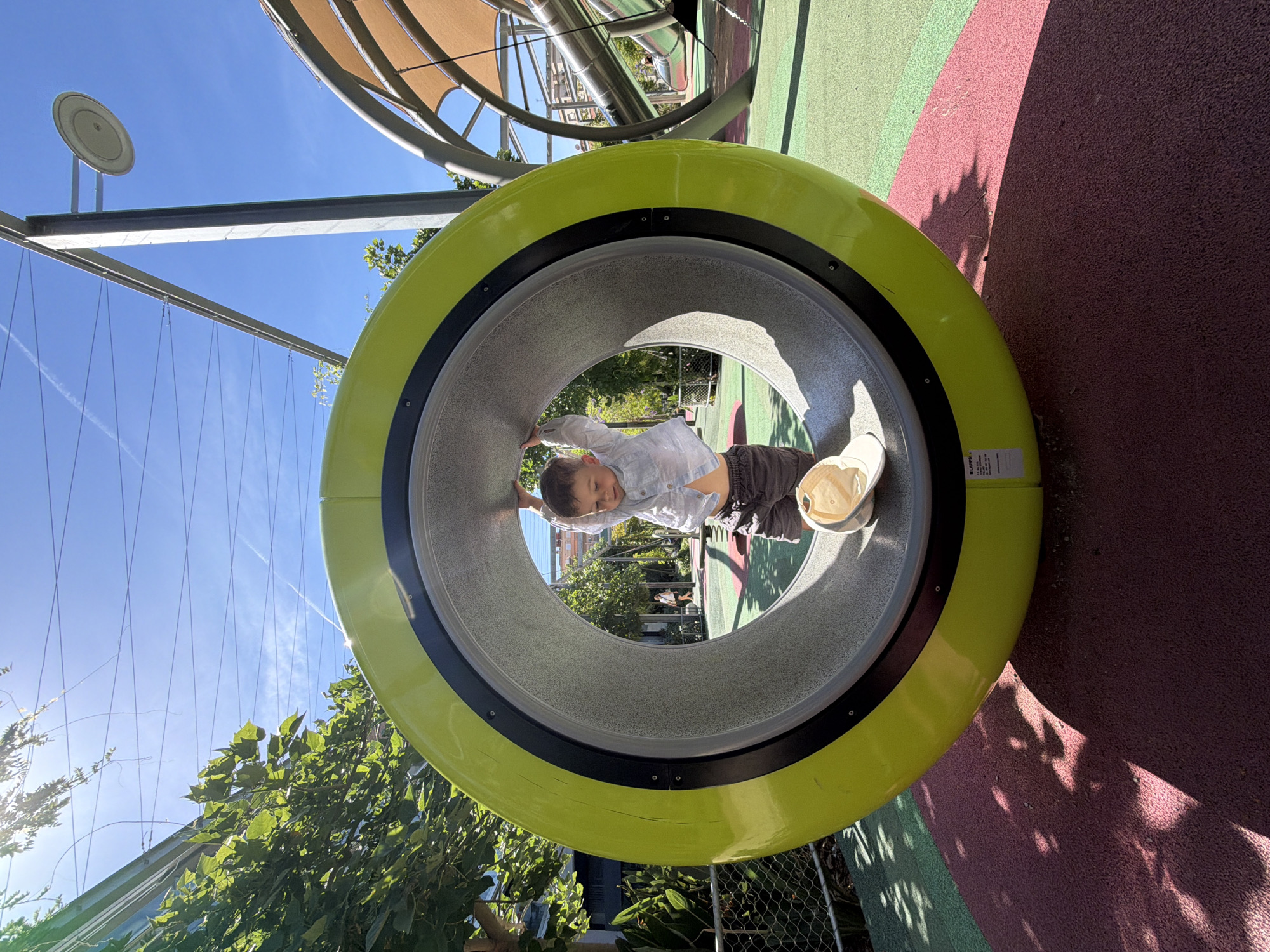 A young child is seen standing inside a large, bright yellow tunnel at a playground. The child, wearing a light blue shirt and grey shorts, is holding onto the inside edge of the tunnel. A light-colored hat rests on the floor of the tunnel. The playground features green and pink rubberized surfaces, a metal structure with a tan canopy, and a street lamp. In the background, trees, overhead wires, and a clear blue sky are visible. A person walks in the distance. The scene appears to be in Barcelona, Spain, suggested by the architectural style and the presence of palm trees often found in the city.