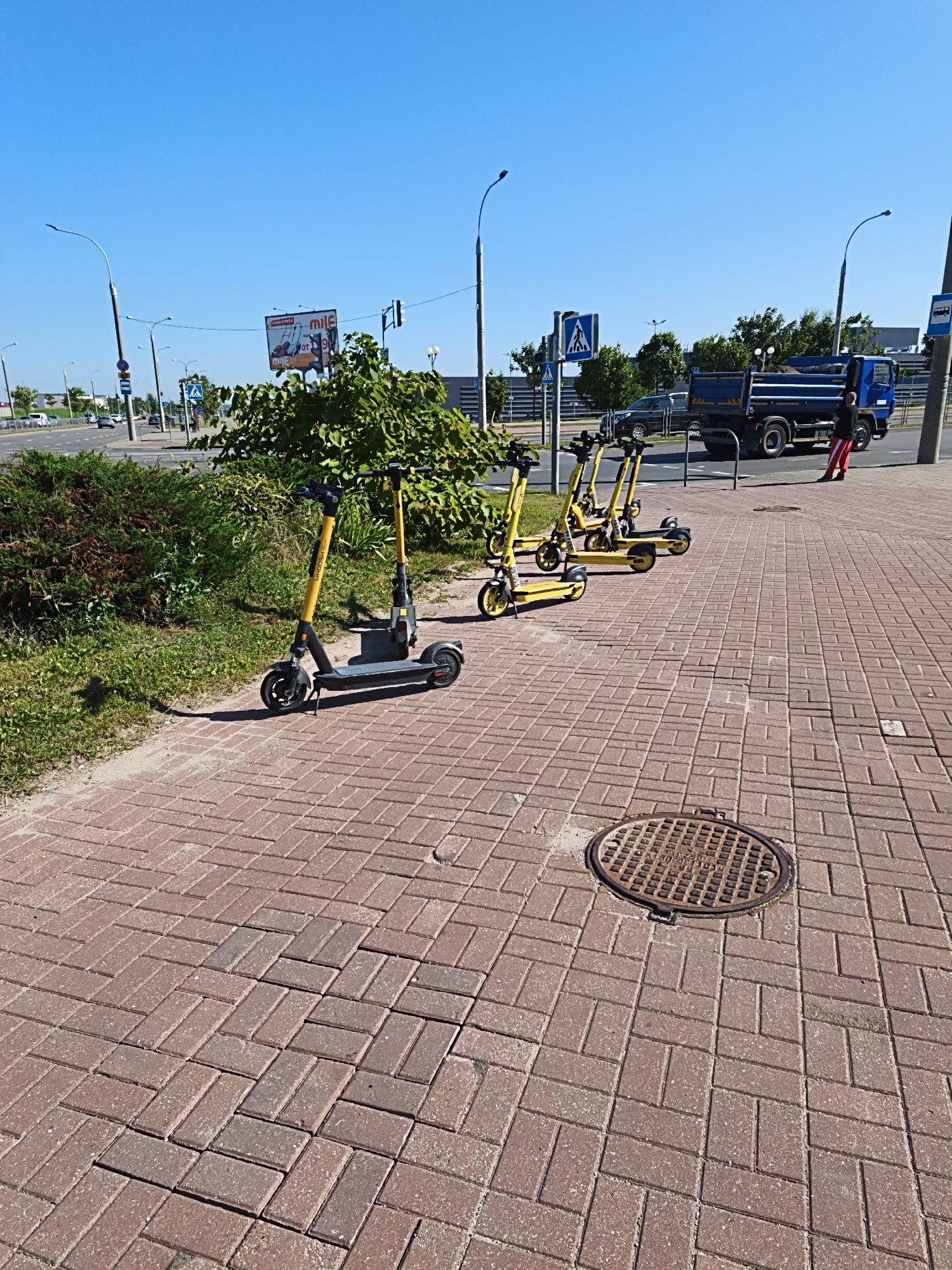 A wide shot captures a sunlit sidewalk in Minsk, Belarus, paved with red bricks laid in a herringbone pattern. Several yellow electric scooters are parked in a row on the sidewalk, their handlebars facing the street. In the background, a blue dump truck is parked on the side of a road, and a person in red pants and a dark shirt stands near it, leaning against a fence. The sky is a clear, bright blue, and green bushes and trees line the edge of the sidewalk and road. A metal manhole cover is visible on the sidewalk in the foreground. A traffic light and street signs are also present in the scene.