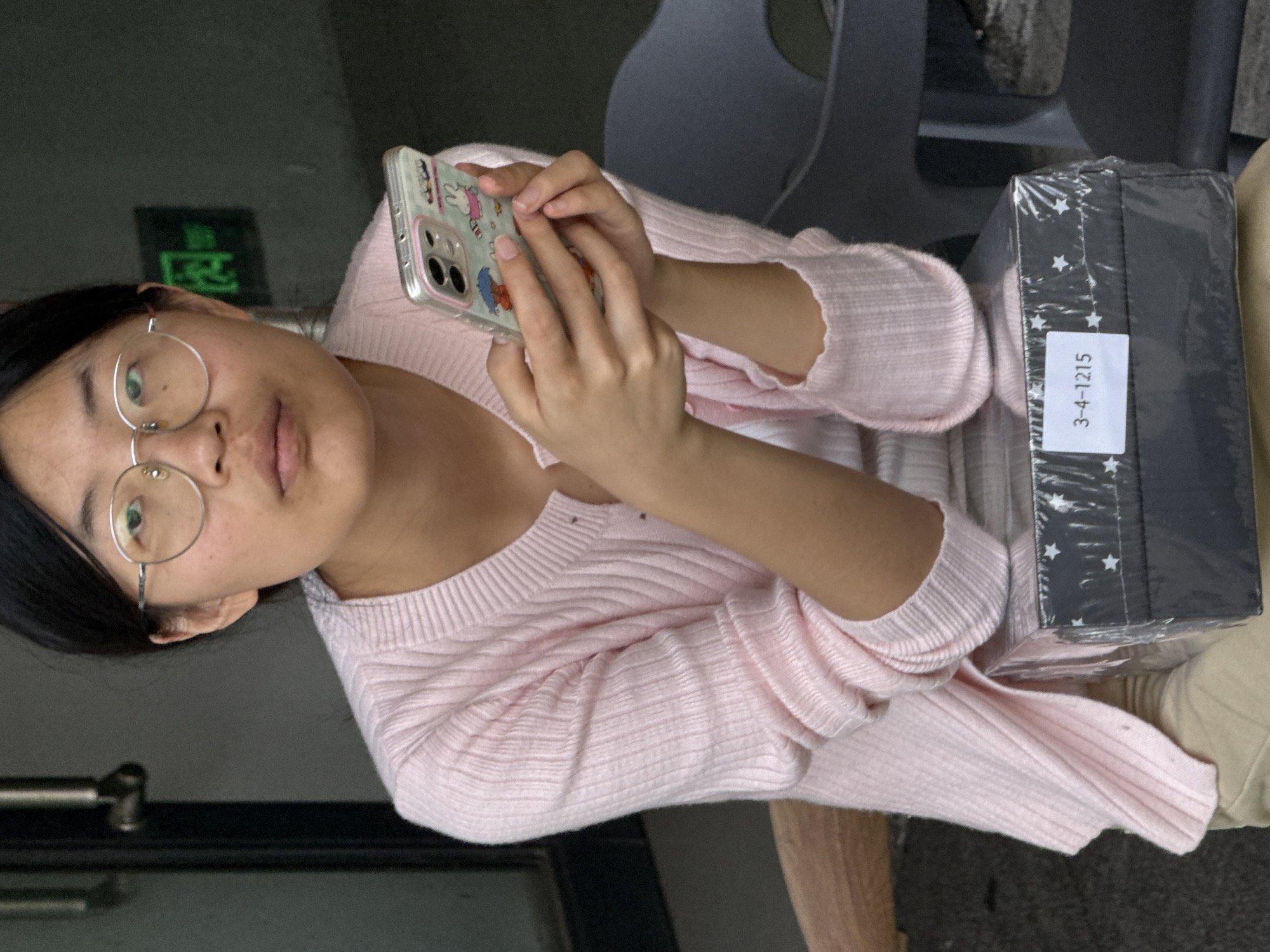 A young woman wearing round-rimmed glasses and a pink sweater sits indoors, holding a smartphone in her hands. A black, rectangular box, wrapped in plastic and labeled "3-4-1215", rests on her lap. The setting appears to be in Jiangjin, China, though precise location cannot be confirmed from the image.