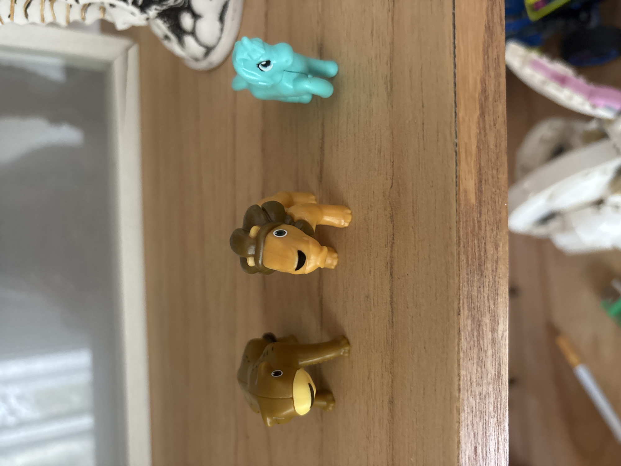 The image shows three small toy animals on a wooden surface. From top to bottom, the toys are a turquoise pony, a brown and tan lion, and a brown elephant. The toys are lying on their sides. The background includes a white picture frame and some other blurry objects. The location is unknown, and there is no information connecting it to Buenos Aires, Argentina.