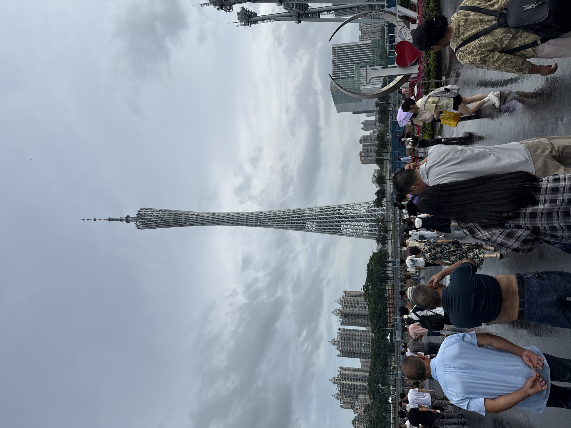 The image depicts a wide outdoor plaza in Tianhe District, China, dominated by the Canton Tower, a tall, lattice-like skyscraper. The plaza is paved with wet, reflective surfaces, indicating recent rain, and is populated by a diverse crowd of people walking in various directions. In the background, a cluster of modern residential buildings and a glass-fronted office building with a distinctive architectural design are visible. To the right, industrial cranes and a large, decorative "LOVE" sculpture with a red heart are present. The sky is overcast with gray clouds. People are dressed in casual attire, and some are carrying umbrellas or shopping bags. The overall scene suggests a public gathering space or tourist attraction.