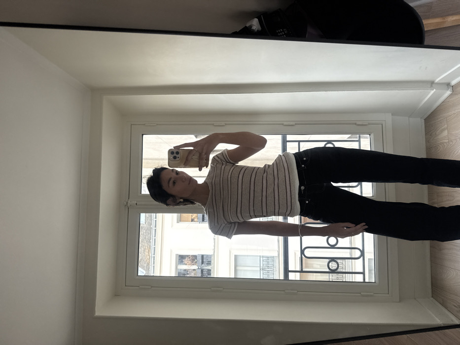 A slender person in black pants and a striped short-sleeved shirt stands in front of a window taking a mirror selfie. The reflection shows the person holding a phone in their right hand. Outside the window, other buildings are visible. A partially visible black chair is in the upper right corner of the mirror. The mirror's frame and a white wall are also visible.