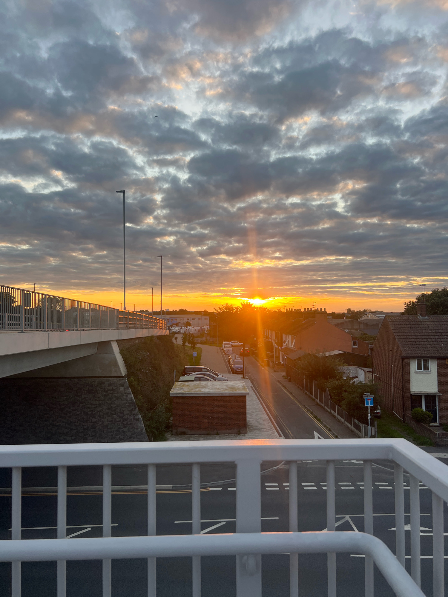 The image captures a sunset over a residential area in Great Yarmouth, United Kingdom. The foreground features a white railing, likely part of a pedestrian bridge or elevated walkway, overlooking a road below. The road has white lane markings and yellow center lines. To the left of the road, a pedestrian bridge with a concrete underside and a stone embankment is visible. Beyond the road, several houses with pitched roofs, some brick and some with white facades, line the street. A few parked cars are visible on the street and in driveways. In the background, commercial buildings and trees can be seen. The sky is filled with dramatic, grey clouds, illuminated by the bright orange and yellow hues of the setting sun, which creates a strong sunburst effect. A single street lamp is visible on the pedestrian bridge. The overall scene depicts a typical suburban environment at dusk.