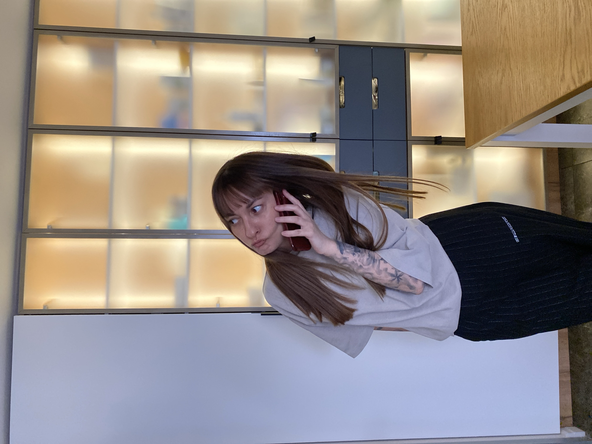 A young woman with shoulder-length brown hair and bangs, clad in a light-colored t-shirt and black striped pants, is holding a red smartphone to her ear, and looking to her left with a suspicious expression. Her left arm is adorned with tattoos. Behind her is a shelving unit with frosted glass doors and metal frames, illuminated from within. To the right, a wooden desk partially occupies the frame. The lower portion of the image is a light gray wall. The city of Votkinsk, Russia cannot be confirmed in this image.