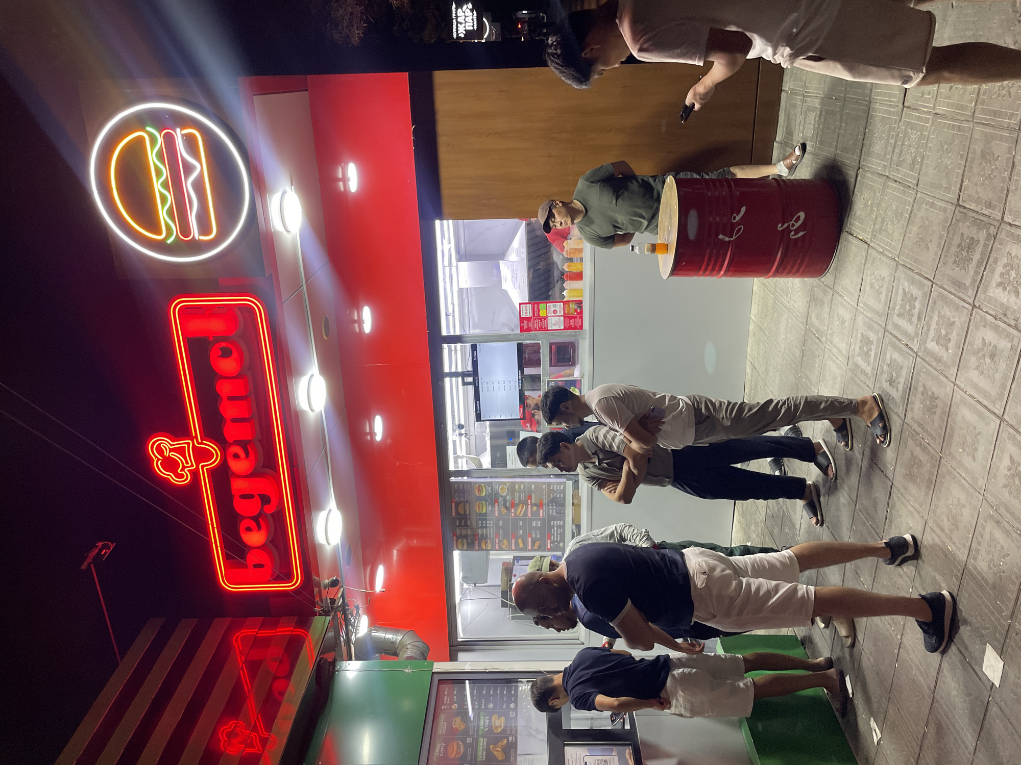 The image depicts a night scene outside a fast-food establishment in Bishkek City, Kyrgyzstan, identified by the prominent red neon sign that reads "begemot" and a stylized burger logo. A group of men are standing in line, seemingly waiting to order food from the counter, which features a menu board. One man in the foreground is looking at his phone, while others are engaged in conversation or observing their surroundings. The establishment itself is characterized by a bright red facade and illuminated windows, suggesting it is open for business. The ground is paved with square tiles, and a distinctive red barrel with white lettering serves as a makeshift table or stand, with a bottle of orange liquid on top. The overall atmosphere is that of a casual, urban night spot.