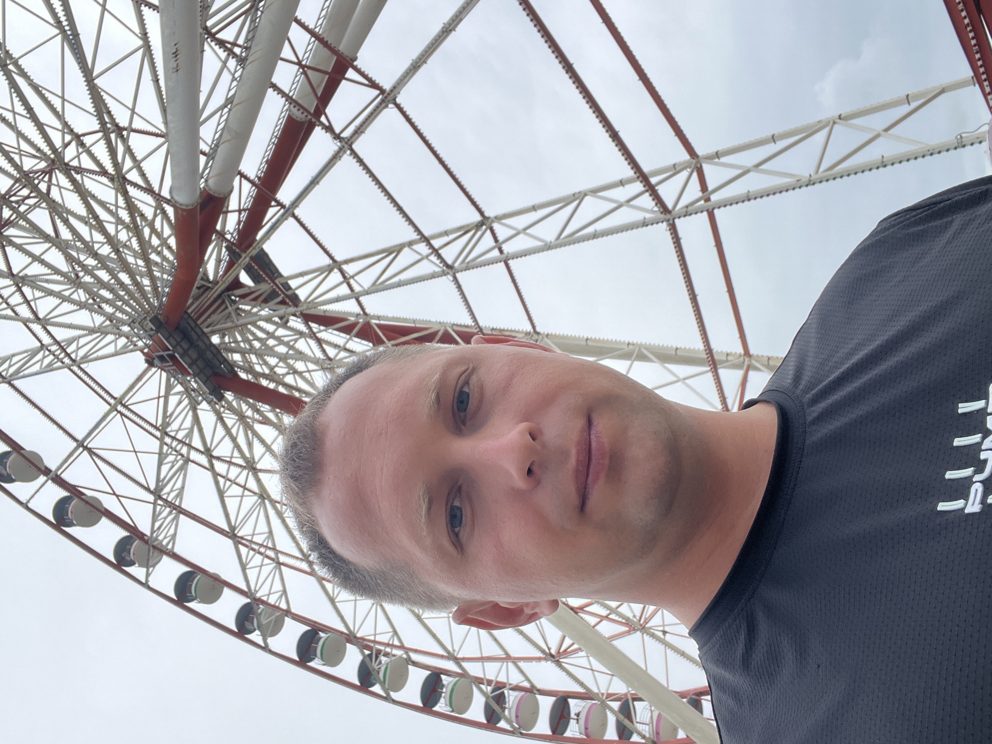A man is taking a selfie in front of the Batumi Ferris Wheel in Batumi, Georgia. The Ferris Wheel is a large attraction with red and white supports and many gondolas. The man is wearing a black t-shirt. The sky is overcast.