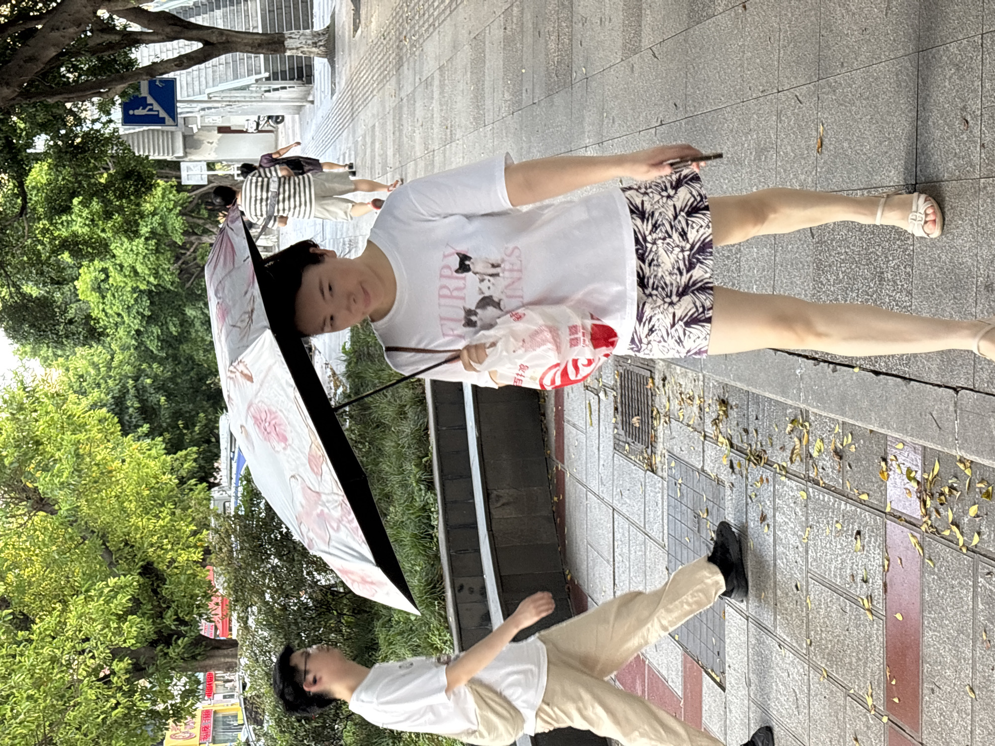 This image shows a street scene in Liangjiang, China. A woman walks on a sidewalk holding an umbrella and a shopping bag. She is wearing a white t-shirt with a cat graphic and a patterned skirt, and light-colored sandals. To her left, a man wearing a white t-shirt, beige pants, and dark shoes walks on the sidewalk. He has a bag slung across his body. In the background, more people are walking and trees line the street. The sidewalk is made of stone and concrete, and fallen leaves are scattered on the ground. A set of stairs with a blue sign indicating a pedestrian pathway is visible to the right.