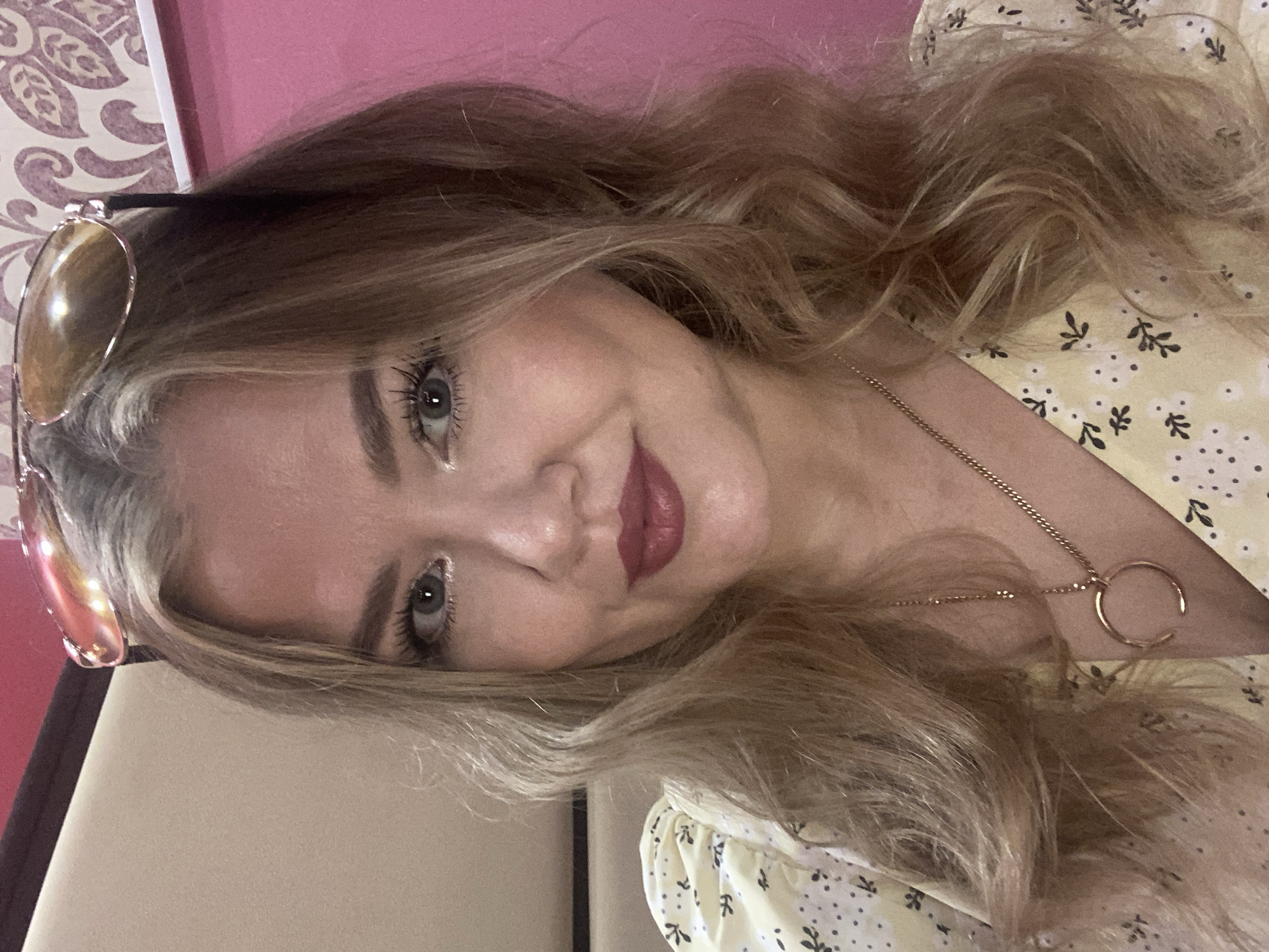 A medium shot shows a Caucasian woman with blonde, wavy hair, wearing a yellow floral-patterned dress and a gold necklace with a crescent-shaped pendant. She is wearing sunglasses on top of her head and has on makeup, including bold lipstick. The woman is smiling, and her head is slightly tilted. The background appears to be an indoor setting with walls of different colors, including pink and a patterned wallpaper. There is no information to suggest this image is located in Yerevan, Armenia.