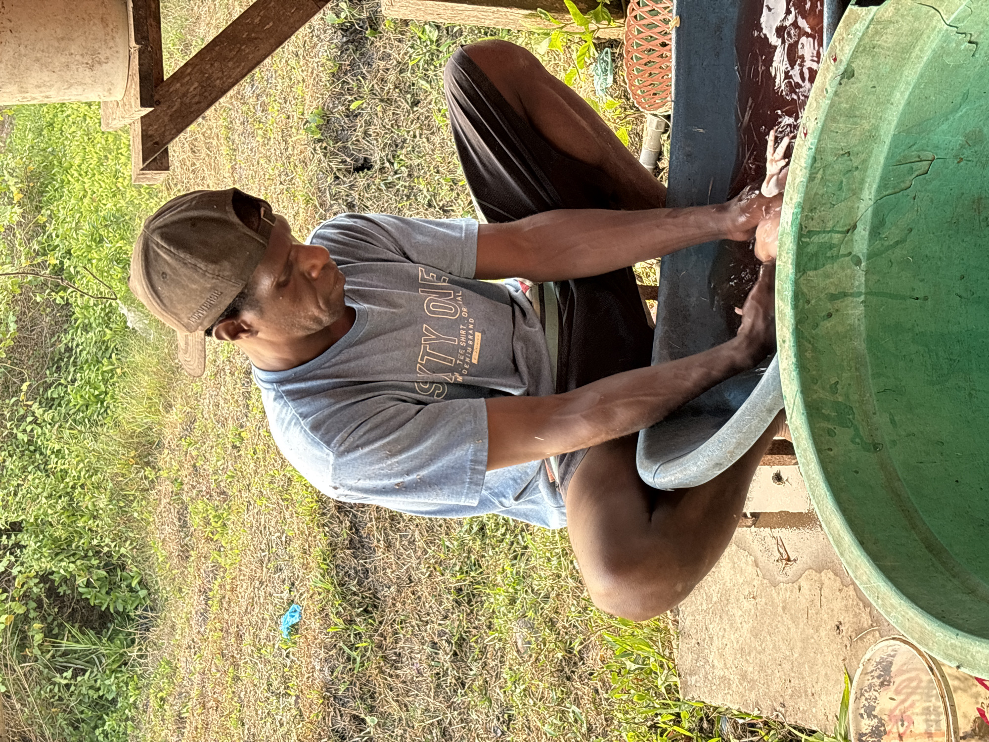 A man wearing a brown baseball cap and a grey t-shirt with text printed on it is squatting next to a blue tub filled with reddish liquid. His hands are in the tub, and it appears he is cleaning something. In front of the blue tub is a large green tub. The background shows dry grass, some green foliage, and a wooden structure with a cylindrical object on it. The scene is outdoors in a rural or semi-rural area. This location is in the city of Merauke, Indonesia.