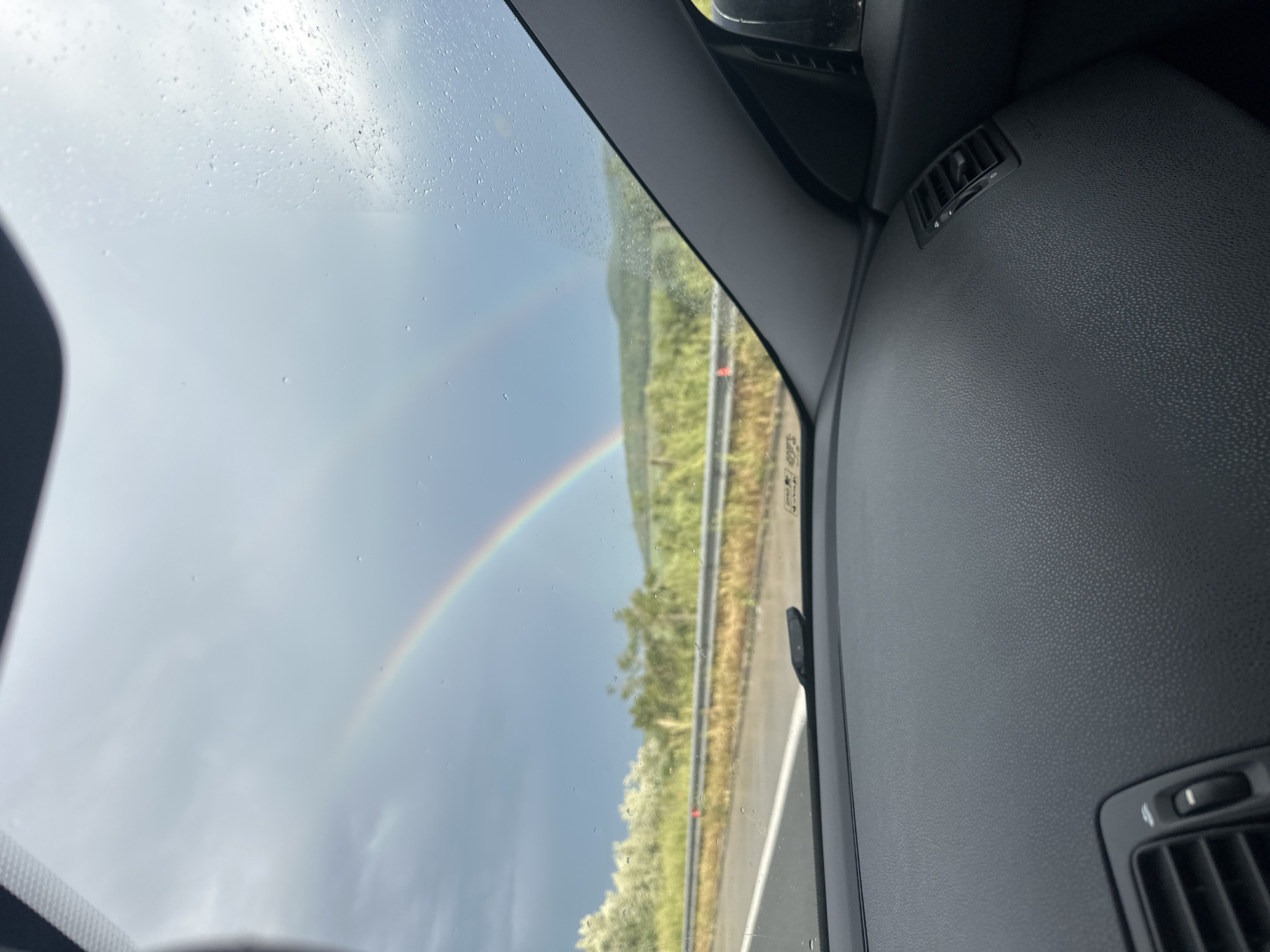 The image, taken from inside a car, shows a double rainbow in the sky. Raindrops are visible on the windshield. Outside, there is a road with vegetation on either side. Part of the car's dashboard is in the foreground. The location is unknown, and there is no indication of people or any action or interaction beyond the car moving through the environment. The image does not provide any information regarding the location in the city of Nevsha, Bulgaria.