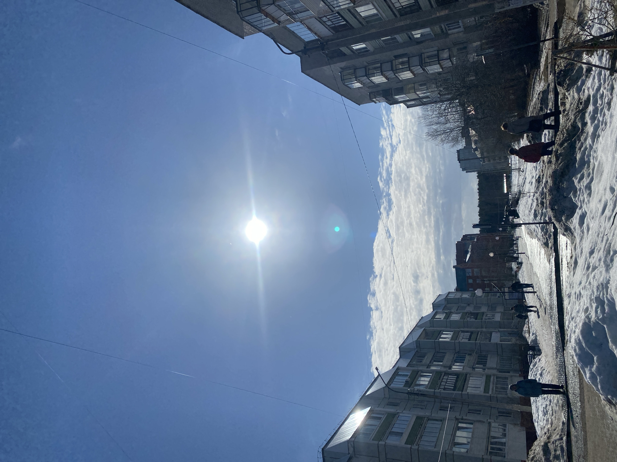 This image captures an urban street scene in the city of Chaikovsky, Russia, observed during a bright, sunny day, likely in late winter or early spring given the snow cover. The sky is predominantly clear blue with scattered white cumulus clouds visible above the horizon, beneath a bright sun positioned centrally in the upper frame, generating a noticeable lens flare. Multiple faint aircraft contrails and overhead power lines crisscross the blue expanse.
On the left, a multi-story residential building features a light-colored facade, numerous rectangular windows, and a prominent sloped, metallic roof. Similar architectural structures extend into the distance along this side of the street. A streetlamp is visible in the mid-ground. On the right, another multi-story residential building, constructed with darker grey bricks, presents multiple bay windows and enclosed balconies.
The ground is characterized by significant accumulations of melting snow, forming irregular piles along the pedestrian pathways. The paths themselves are partially cleared, exposing wet, reflective pavement or ice. Several individuals are present: one person wearing a blue-grey jacket stands on the path in the left foreground. Further along the left path, additional figures are visible walking. On the right, two individuals are walking away from the viewer; one wears a red jacket, and the other a light-colored coat. Bare deciduous trees and shrubs are interspersed between the buildings and extend into the background, where more distant urban structures and a dark line of trees or forest are visible.