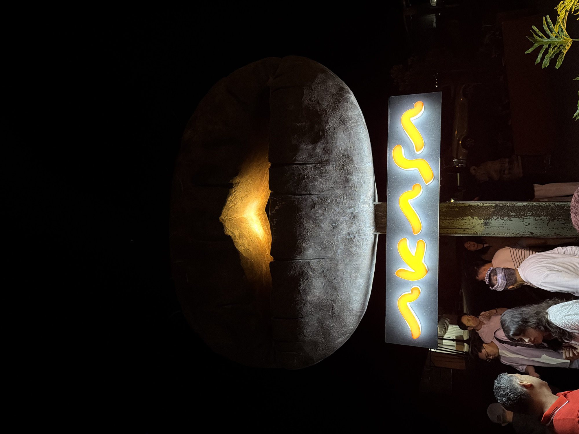 In Kintamani, Indonesia, at night, a sign with illuminated orange script that reads "AKASA" is affixed to a pole. The sign is positioned below a large, dark, circular sculpture that glows from within, emitting a warm orange light. A group of people are gathered in front of the sign, some standing and talking, while others appear to be entering or exiting a building to the right. The scene is dimly lit, with the primary light sources being the sculpture and the sign itself. The background is dark and indistinct, with some foliage visible in the foreground.