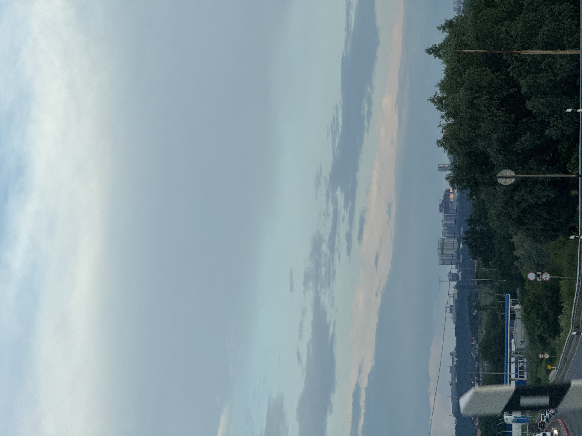 The image captures a panoramic view of the outskirts of Novomar'evskaya, Russia. The foreground features a highway with a white and black striped traffic delineator post on the left. A few cars are visible on the highway. In the middle ground, there is a gas station with a blue canopy, surrounded by lush green trees. Further in the background, a dense line of trees forms a natural barrier before a distant cityscape unfolds. The cityscape is characterized by numerous apartment buildings and construction cranes, indicating ongoing development. The sky above is a blend of soft blue and grey clouds, suggesting either early morning or late evening light. The overall scene depicts a transition zone between a rural highway and an expanding urban area.
