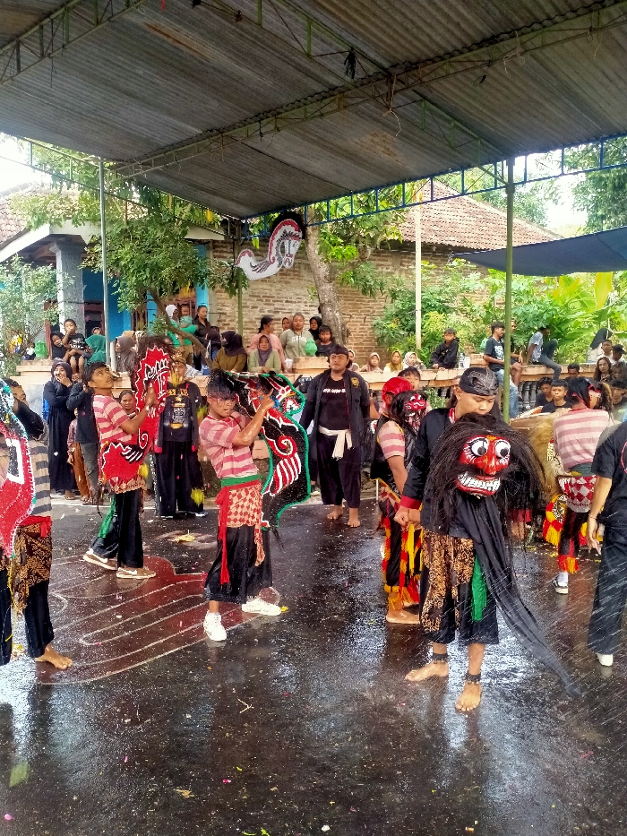 The image depicts an outdoor cultural performance in Jenggrik, Indonesia, under a corrugated metal roof structure supported by green trusses. The ground is dark, wet, and reflective, with white chalk-like markings and scattered debris.
In the mid-ground and foreground, approximately five to seven individuals, primarily male, are engaged in a performance. Two younger performers, centered, wear red and white striped shirts, black pants, and patterned red/white/green sarongs or sashes. Each holds a large, brightly colored, intricately patterned flat horse effigy (kuda lumping). The performer on the left wears white sneakers, while the one on the right appears barefoot. To the right, another performer wears a black top, patterned skirt, and a prominent red-faced mask with exaggerated white eyes, a wide mouth, and black hair-like adornments, along with a long black fabric extension. This performer is barefoot with dark ankle adornments. Other performers in traditional attire are partially visible. A man in a black jacket and black pants stands behind the central performers.
In the background, a diverse crowd of onlookers, including adults and children, is observing the event. They are standing, sitting on benches, or leaning against brick walls and structures. Behind the crowd, various residential buildings with tiled roofs, brick walls, and lush green foliage are visible. A white, stylized horse figure is mounted on a brick wall to the left of center. A large blue tarp or awning is visible on the far right. The overall lighting is diffuse, consistent with an overcast sky.