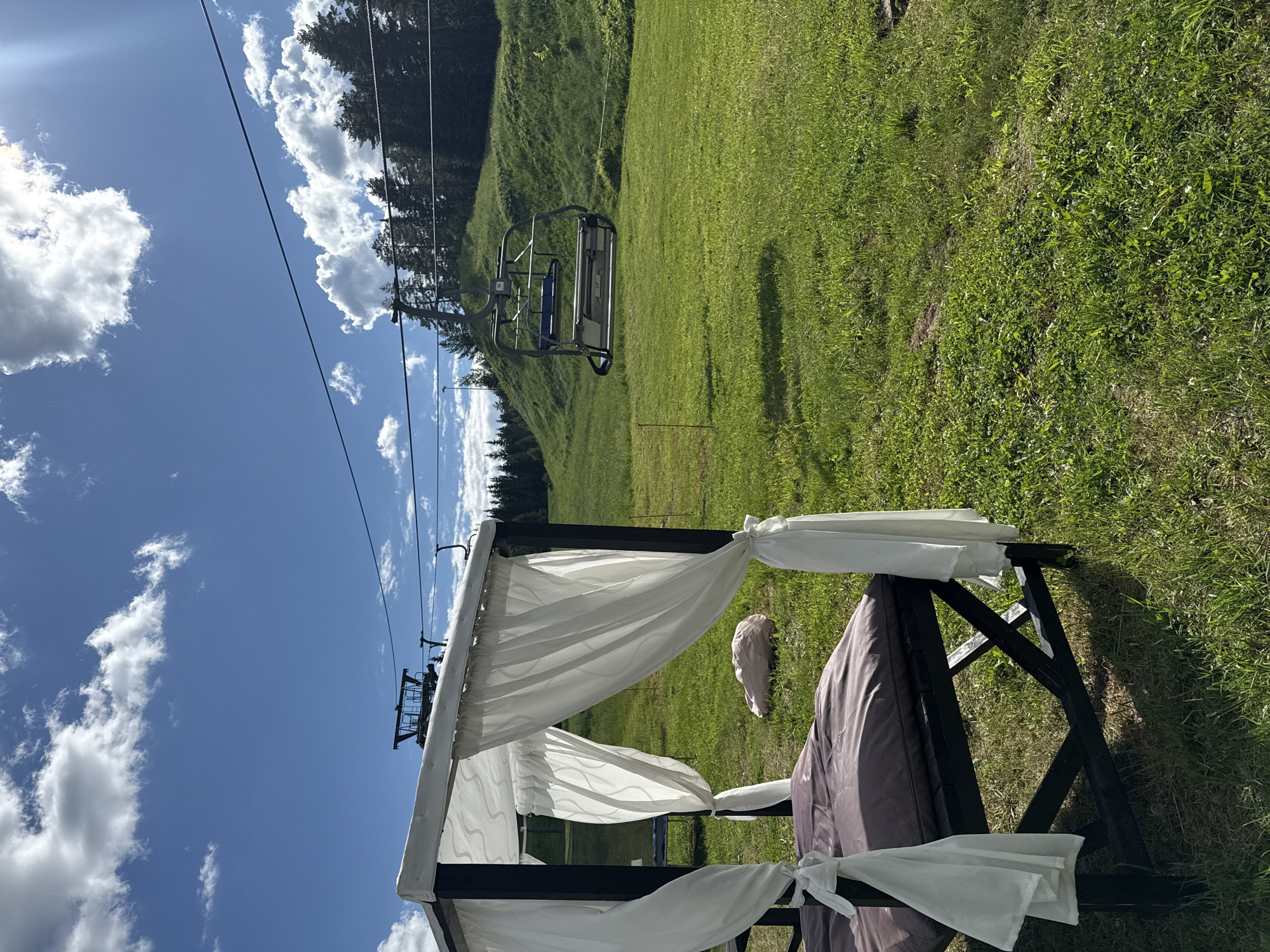 An outdoor scene on a sunny day features a gazebo with white curtains draped around it. A dark brown mattress is placed inside the gazebo. In the background, a ski lift with chairs is visible, suggesting the location is likely a ski resort or a mountain area, possibly in the city of Sheregesh, Russia, during the summer season. The ground is covered in lush green grass, and a few scattered clouds adorn the bright blue sky. A single pinkish-beige rock rests on the grass behind the gazebo.