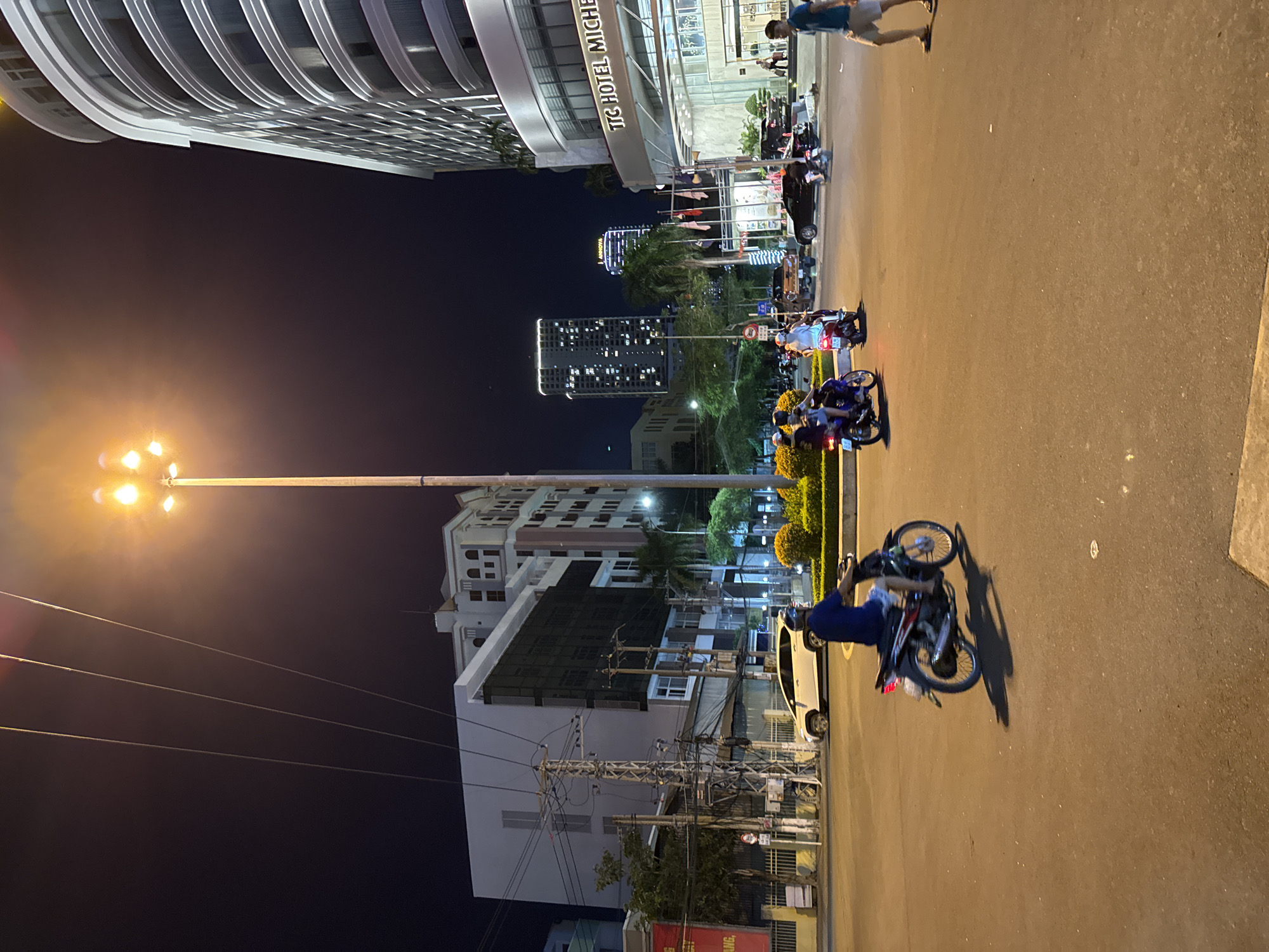 The image depicts a street scene at night in Nha Trang Ward, Vietnam. A prominent modern building with curved architecture and illuminated windows, bearing the text "TG HOTEL MICHE", dominates the left side of the frame. In the foreground, a streetlamp with multiple bright bulbs illuminates the asphalt road. Several people are visible, most of whom are riding motorcycles. One person is standing on the sidewalk near the hotel, while others are on their motorcycles, some appearing to be in motion. The lighting suggests it is nighttime, with the artificial lights of the buildings and streetlamp casting a warm glow on the surroundings. The scene is bustling with activity, indicated by the presence of multiple vehicles and people. The overall atmosphere seems lively, typical of a commercial or residential area at night. The image captures a slice of urban life in Vietnam, with the modern architecture contrasting with the common mode of transportation.