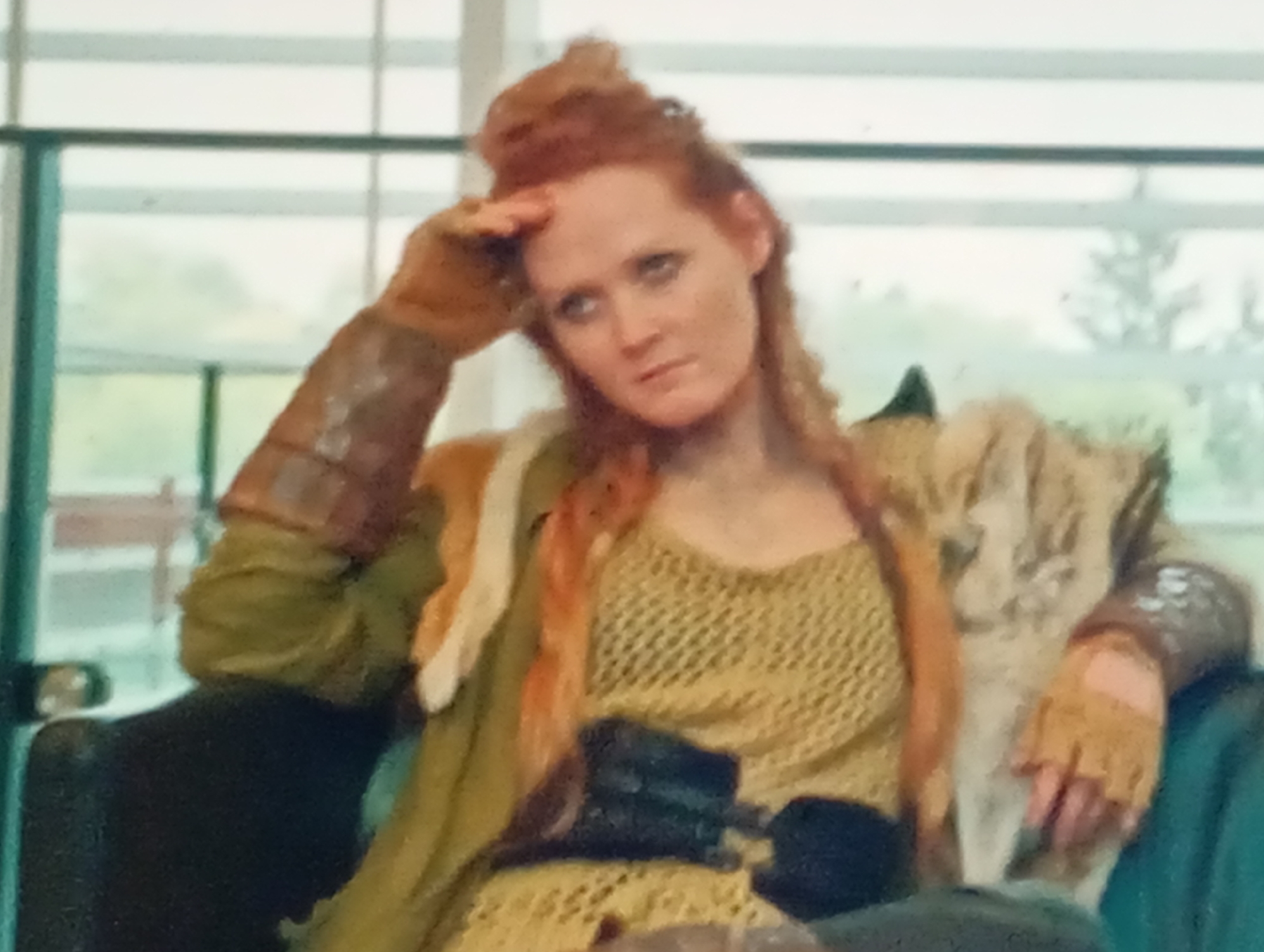 A medium shot captures a woman with red hair styled in an updo, her hand raised to her forehead as if shielding her eyes from the sun or in a contemplative gesture. She appears to be sitting in an indoor setting, possibly a waiting area or a casual lounge, with large windows in the background that suggest an outdoor view. The background outside the windows is blurred, showing trees and a cloudy, overcast sky, hinting at daytime.
The woman is dressed in a rustic, possibly historical or fantasy-inspired costume. She wears a textured, olive-green garment layered with a lighter, possibly fur or fabric draped over her shoulder. Beneath this, she has on a mustard-yellow, netted top, and a dark belt cinches her waist. Her arms are adorned with protective armguards made of leather or similar material, with one on her right arm featuring intricate detailing. Her left hand, also gloved, rests on her lap. Her expression is one of mild weariness or introspection, with her gaze directed slightly downwards and to the side. The overall color palette is earthy, with dominant tones of green, yellow, and brown. There are no visible signs of text in the image. The scene evokes a sense of pause or reflection for the subject.