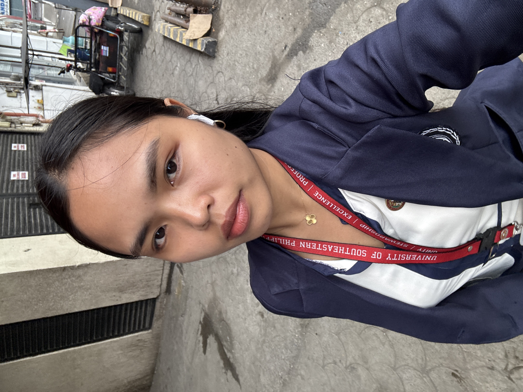 A young woman with dark hair pulled back is taking a selfie outdoors. She is wearing a navy blue blazer over a white top with blue trim and a red lanyard with text that reads "UNIVERSITY OF SOUTHEASTERN PHILIPPINES" and "EXCELLENCE | STEWARDSHIP | PROFESSION". She has a small gold necklace with a clover-shaped pendant. An earbud is visible in her right ear. In the background, there is a sidewalk, a concrete wall with horizontal vents, and some pipes and construction materials. Further in the background, a three-wheeled vehicle with a canopy is parked. The scene appears to be an urban environment, possibly near a school or institution.