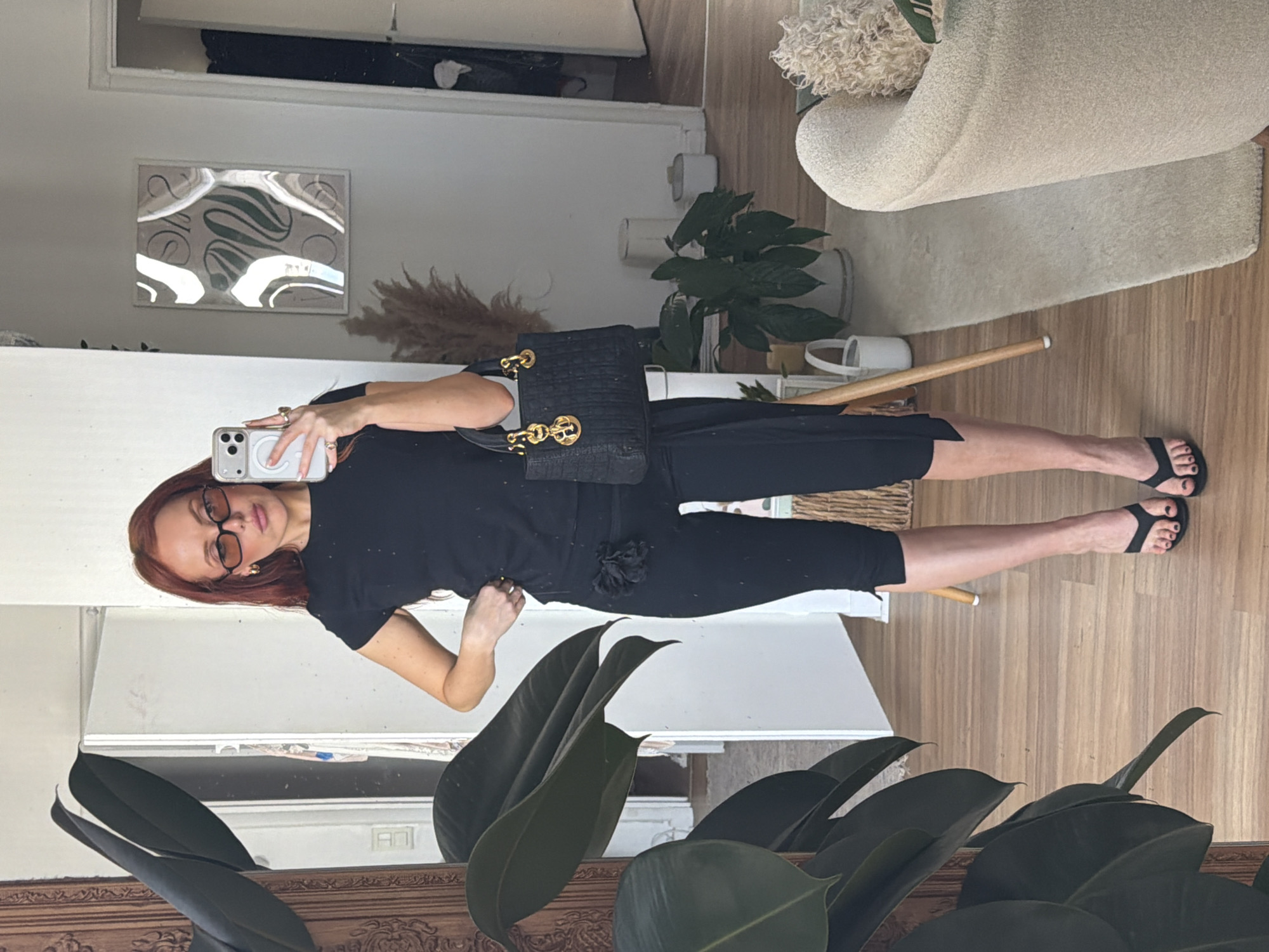 A woman is taking a selfie in a room that appears to be a Parisian apartment. She is wearing a black t-shirt, black shorts, and black sandals, and is carrying a black handbag with gold hardware. She has red hair and is wearing sunglasses. The room is decorated in a minimalist style with white walls, light wood floors, and a cream-colored chair. There is a plant in the foreground and another plant in the background. There is also a picture on the wall. The mirror on the wall reflects a partial view of the interior of the apartment.