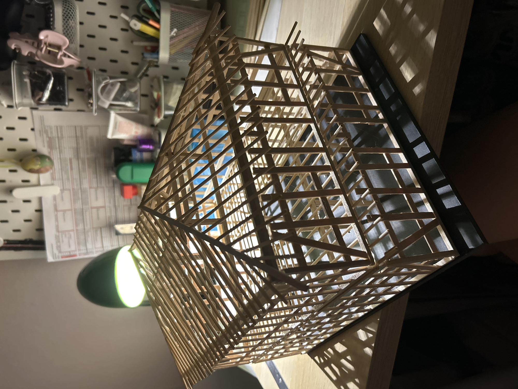 The image depicts a detailed architectural model of a wooden structure, possibly a house or a barn, constructed from thin wooden dowels and planks. The model features a multi-level frame with visible internal supports and a pitched roof structure. It rests on a dark base, which is placed on a wooden surface, likely a desk or table. The scene is illuminated by a green desk lamp positioned behind and to the left of the model, casting strong shadows. In the background, a pegboard organizer holds various supplies, including tubes, containers, clips, and writing utensils, suggesting a crafting or study environment. There are no people or animals visible in the image. The provided location of Sofia, Bulgaria, cannot be visually confirmed from the image content.