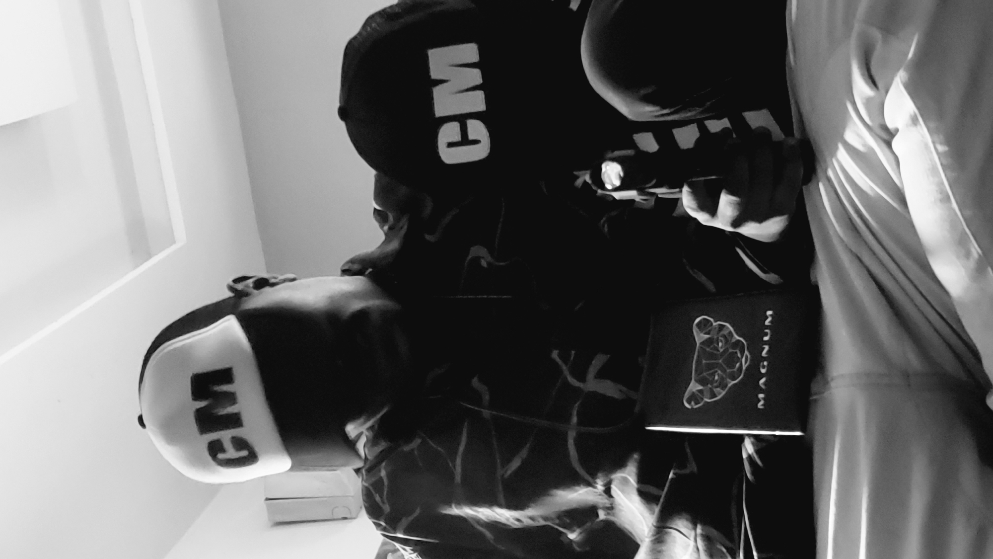In a dimly lit indoor setting, possibly a bedroom in Narayi, Nigeria, a person wearing a patterned shirt and a "CM" branded cap is holding a handgun. Another "CM" branded cap rests on their shoulder. In front of the person, a dark-colored booklet with a geometric animal face design and the word "MAGNUM" is visible. The person is seated, and their lower body is covered by light-colored fabric, possibly bedding. The overall tone of the image is dark and gritty, with strong contrasts between light and shadow.