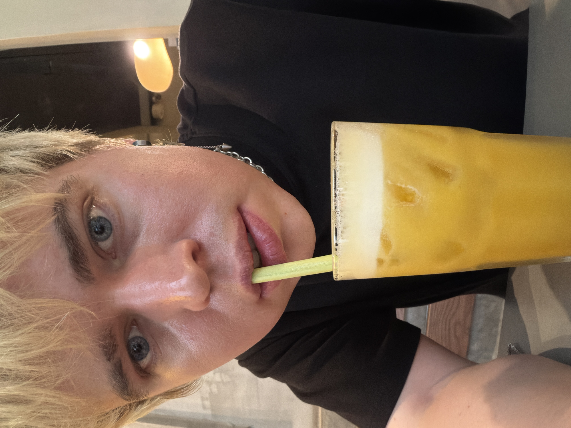 This full-frame, eye-level, selfie-style photograph, taken in Thủ Đức, Vietnam, captures an individual with fair skin, short blonde hair, and blue eyes looking directly at the camera. The person, positioned centrally, is wearing a black short-sleeved crew-neck top. A silver chain necklace with small, dark, conical pendants hangs around the neck, with a dark stud earring visible in the left earlobe. The individual is in the process of drinking from a tall, clear glass containing a vibrant yellow-orange beverage through a light green straw held between the lips. The drink has ice cubes and a discernible layer of white foam at the top. The background is softly blurred: to the left, a light-colored, possibly textured wall or surface is visible; to the upper right, a warm, orange-glowing light fixture is present against a dark background, suggesting an indoor setting like a cafe or restaurant. Below the light fixture, dark fabric or upholstery is visible, along with horizontal wooden planks further down. The person's left arm and a grey table surface are visible in the lower left foreground.